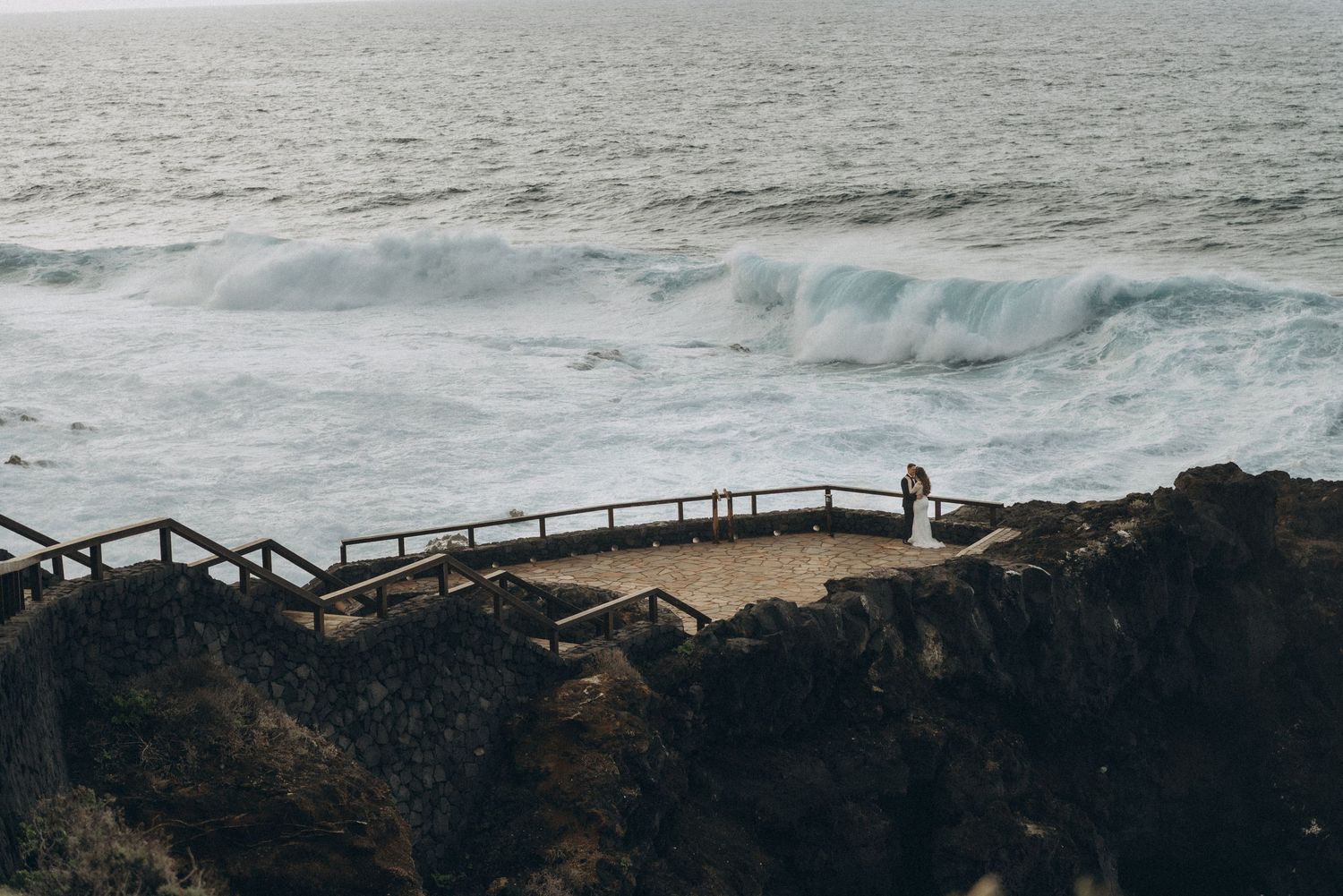 Dramatic aerial view of waves crashing against rocky cliffs with wooden walkways along the rugged coastline. Wedding photoshoot.