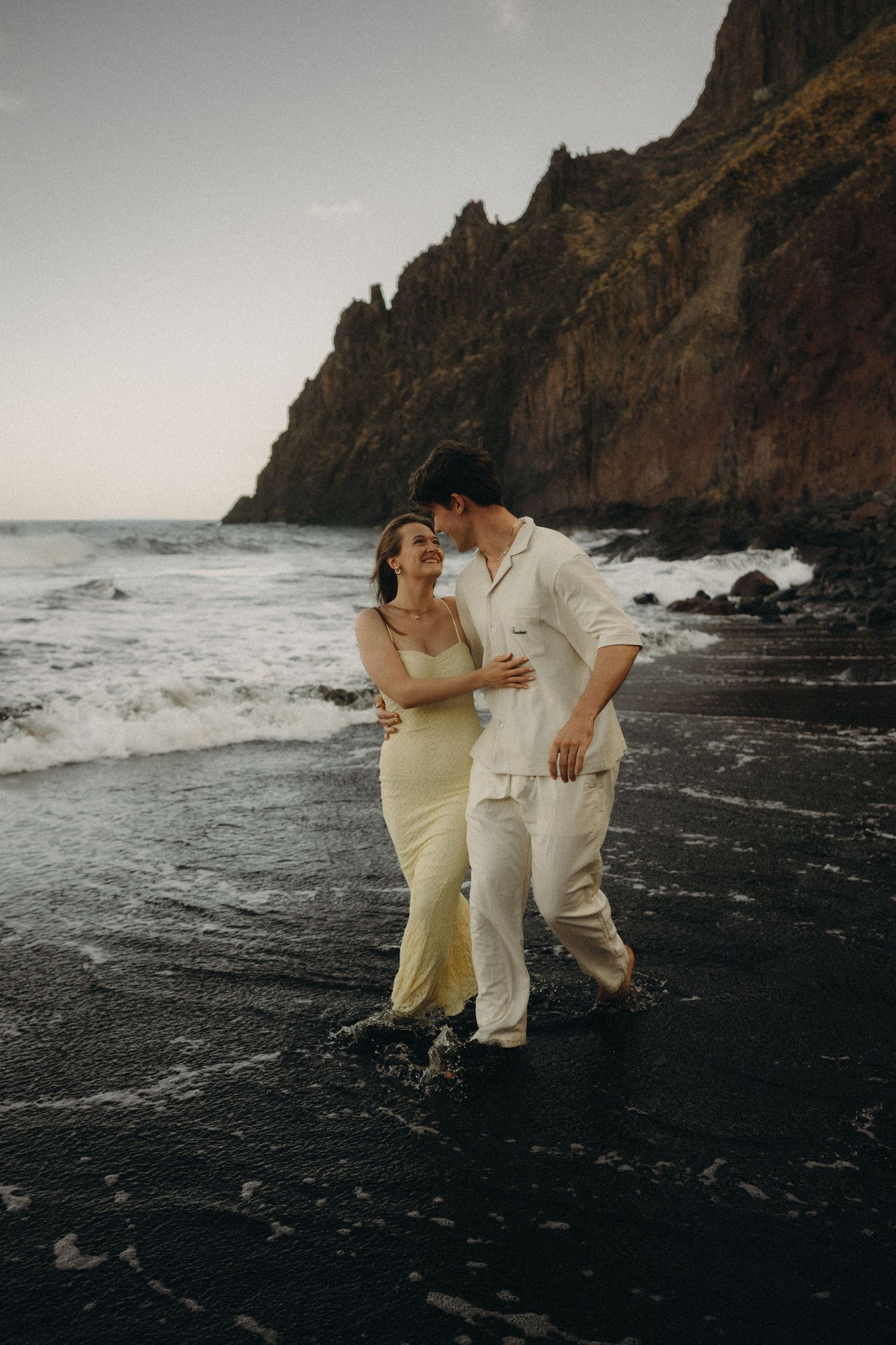 Couple walking on black sand beach at sunset, woman in yellow dress, man in white outfit, rocky cliffs in background.