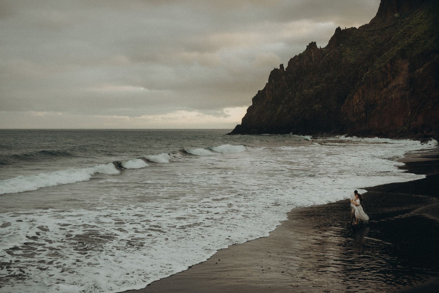Person standing on a dark volcanic beach with dramatic ocean waves and misty cliffs under an overcast sky.