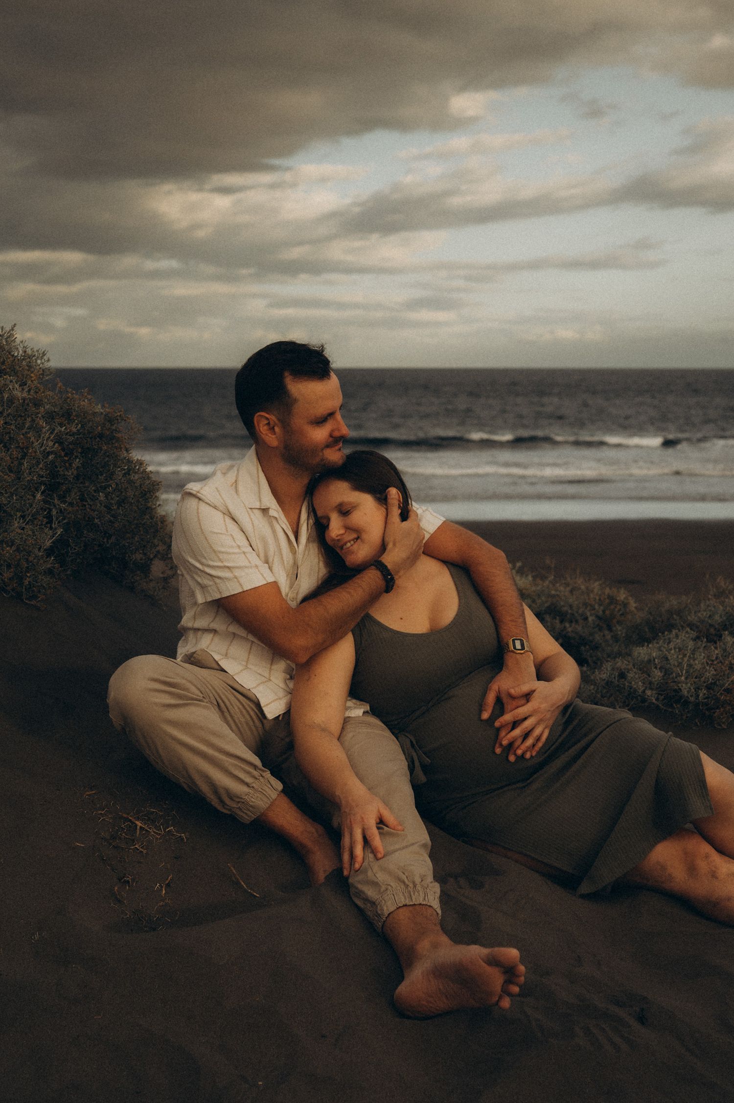 Couple sitting together on a sandy beach at sunset, woman with baby bump leaning into man's embrace by the ocean.