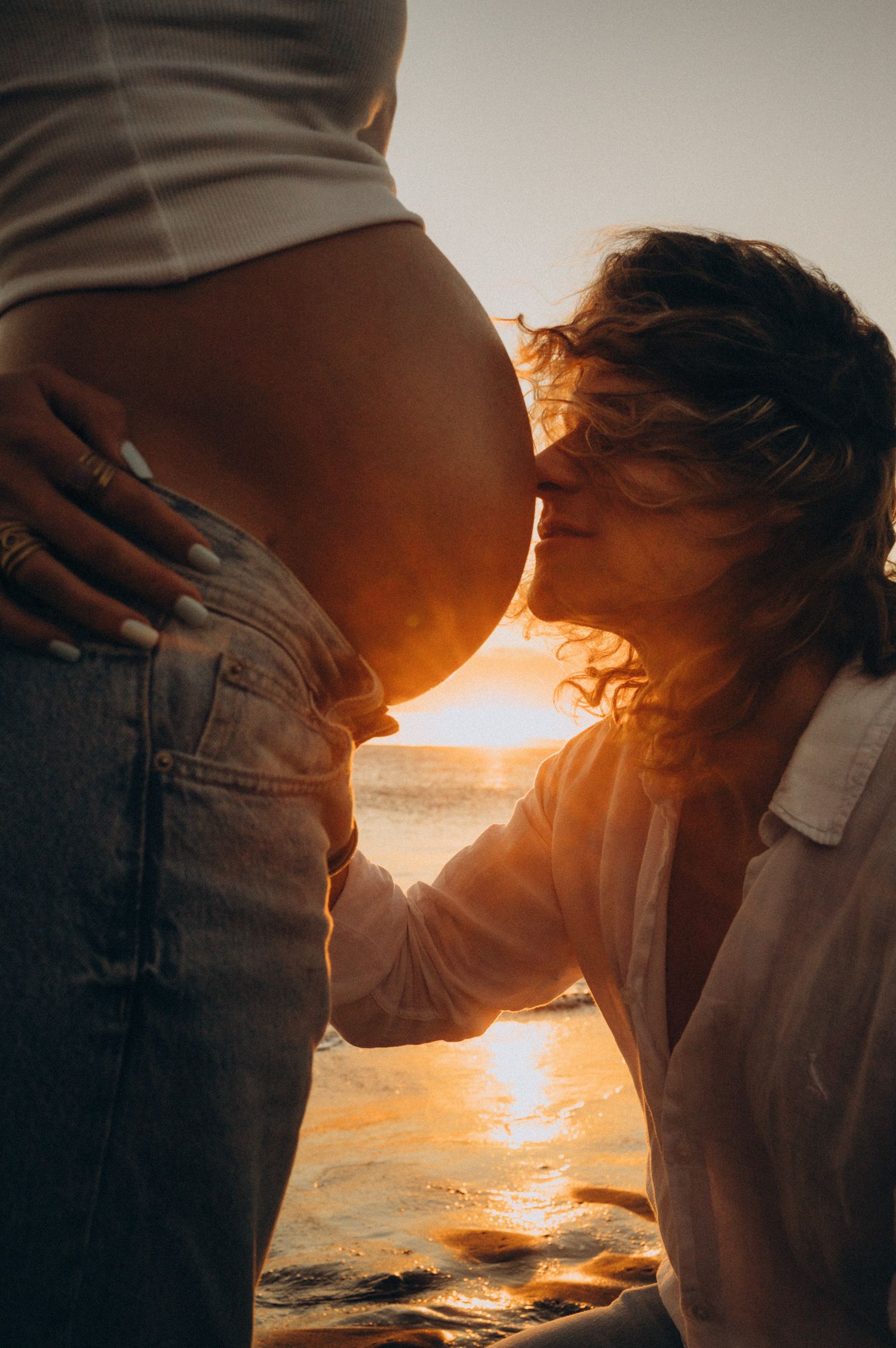 Partner kissing pregnant belly at golden sunset on beach, romantic maternity photo in warm light.