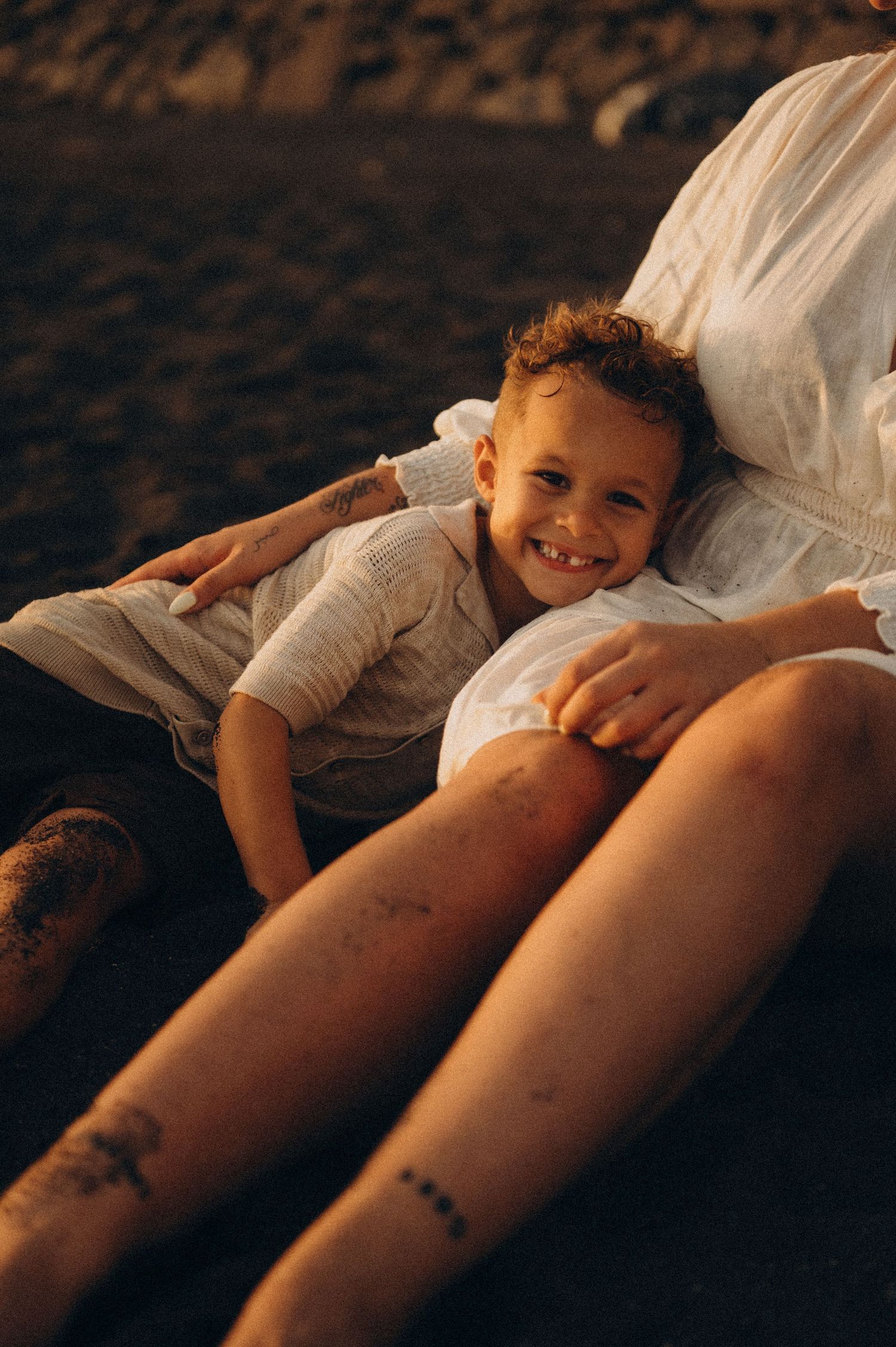 Close-up of smiling toddler in mother’s arms sitting on warm dark sand at sunset, Playa del Duque