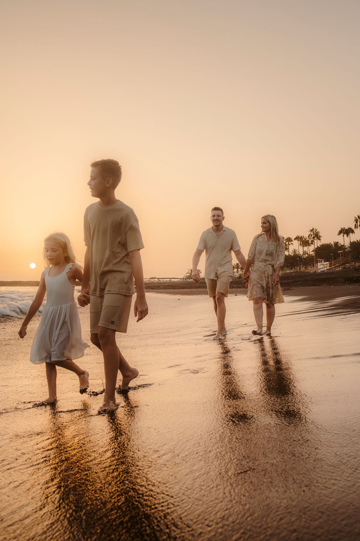 Children holding hands walking along beach with parents behind at sunset, reflections in sand