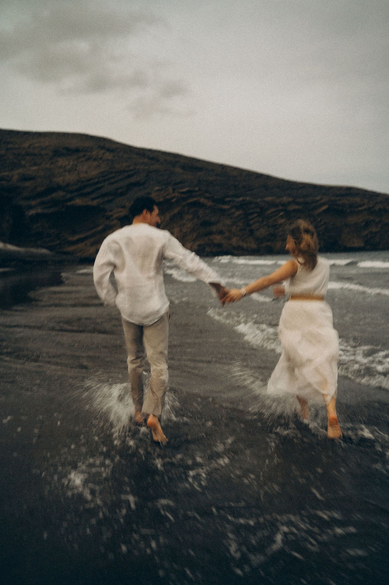 Couple holding hands running through ocean waves on a dark volcanic sand beach with moody overcast skies.