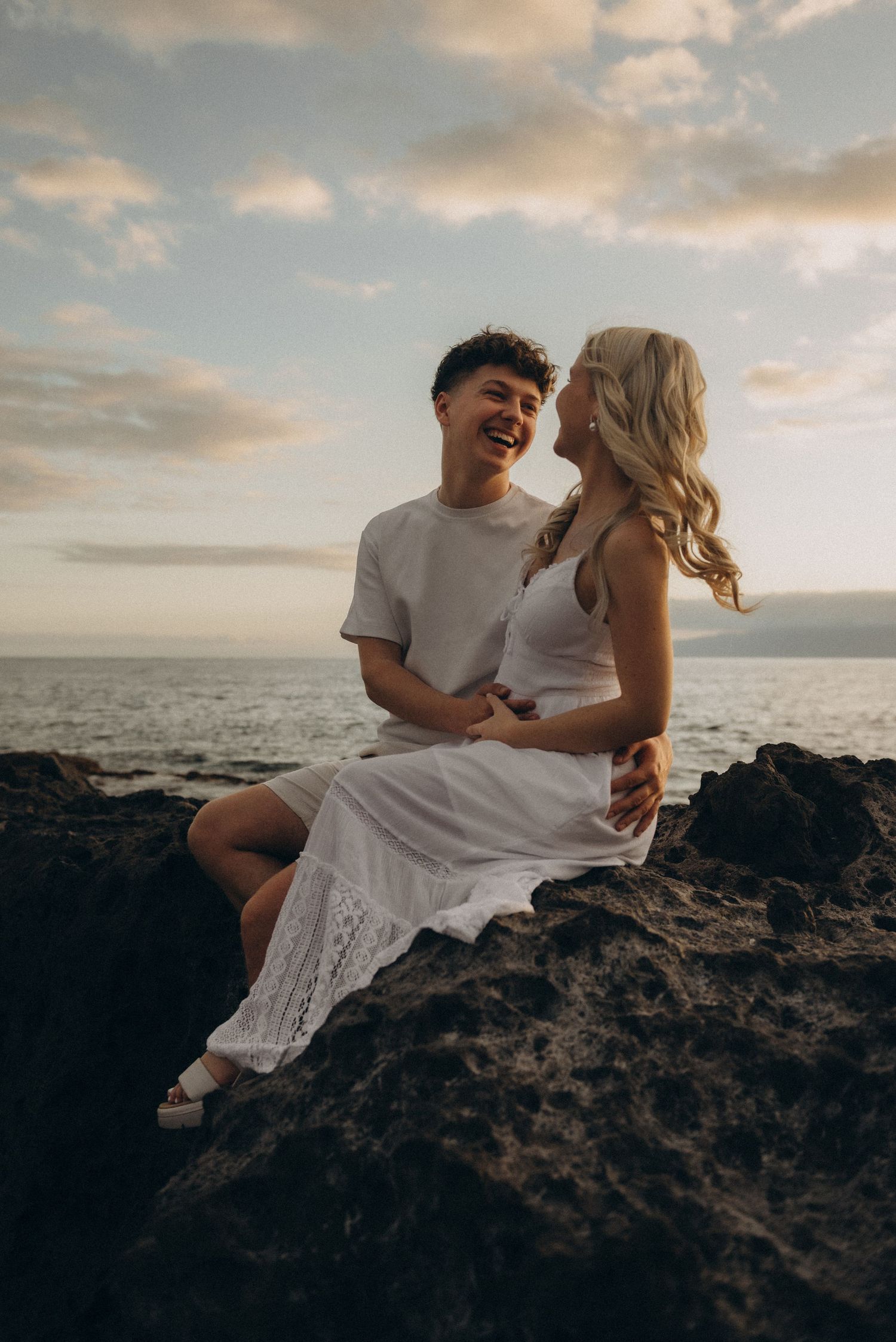 Couple in white outfits laughing on rocky ocean cliffs at sunset.