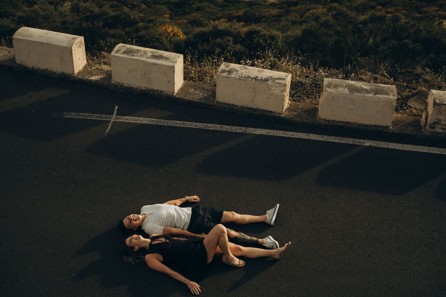 Two people lying on dark asphalt road near concrete blocks at dusk.