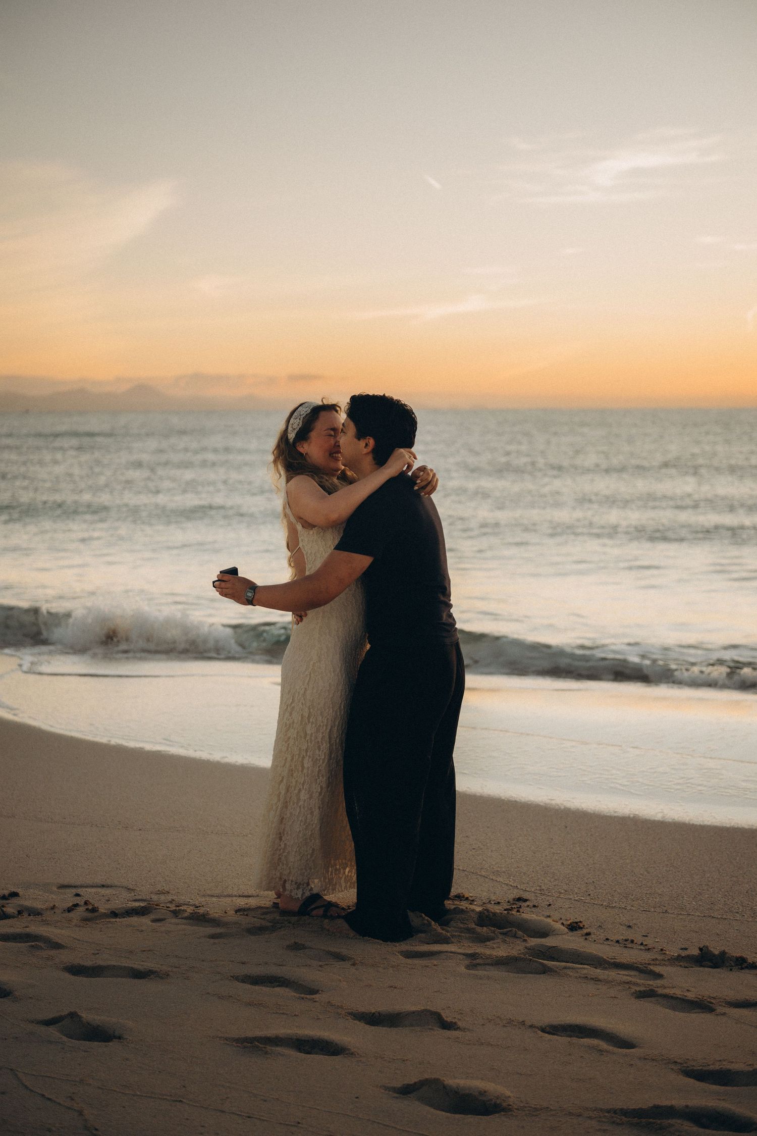 Couple sharing a romantic kiss on a sandy beach at sunset with ocean waves in the background.