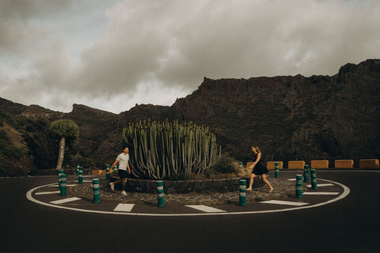 Couple walking around a circular roundabout with a large cactus centerpiece, surrounded by mountains under cloudy skies.