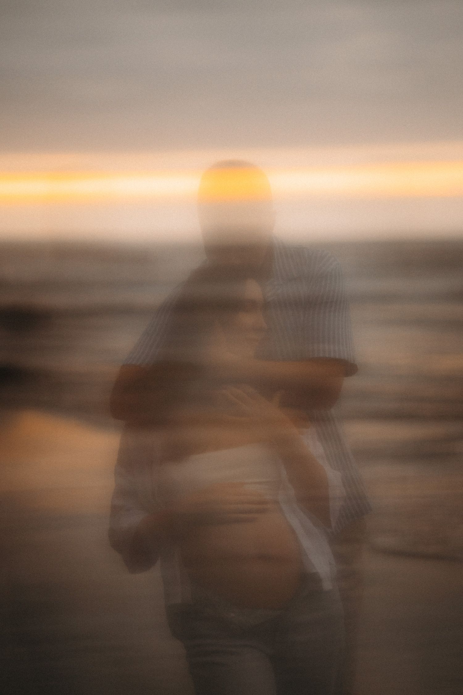 Blurry artistic photo of a pregnant couple embracing on a beach at golden sunset.