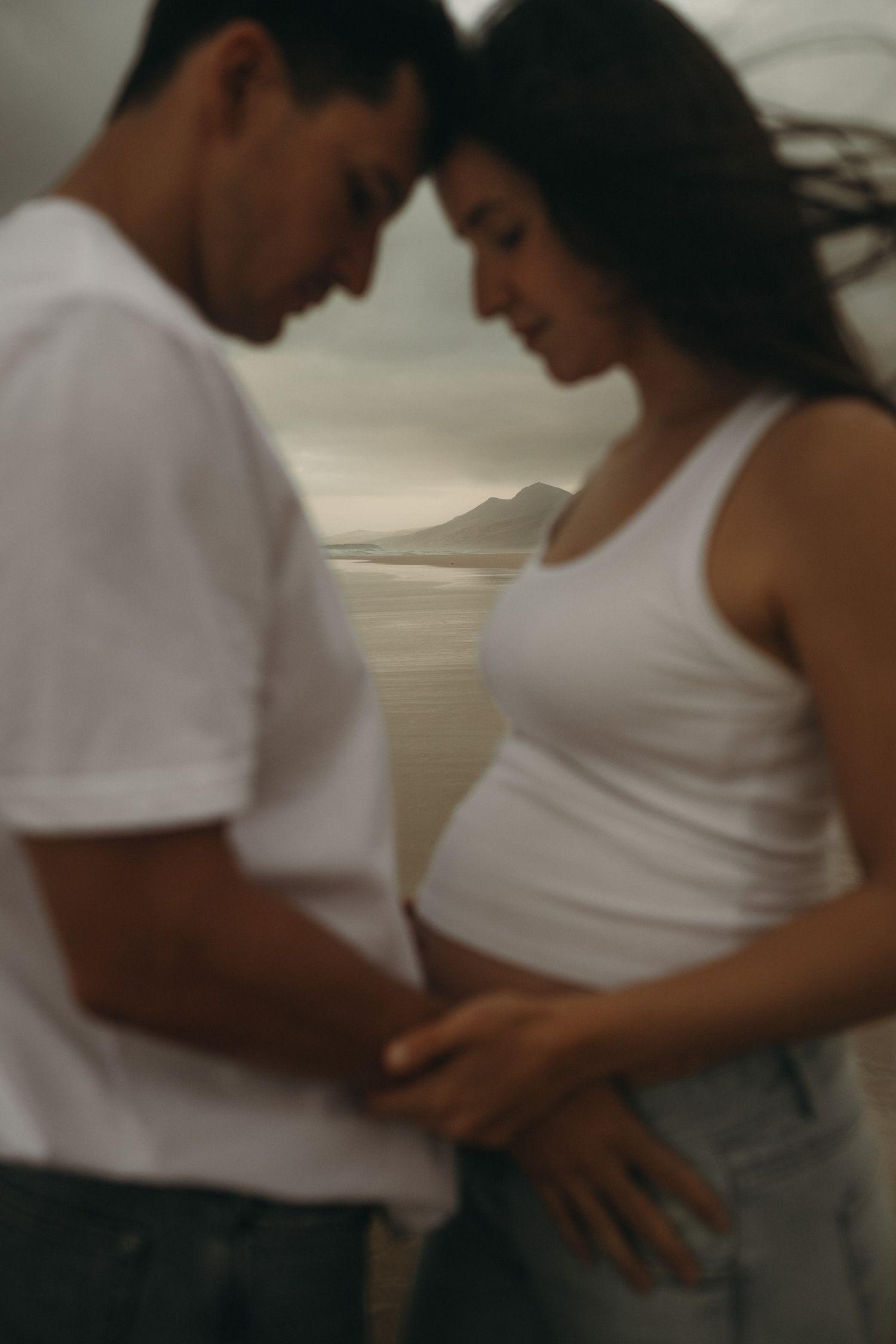 Couple tenderly touching pregnant belly on beach with mountain backdrop, wearing white tops in an intimate maternity photo.