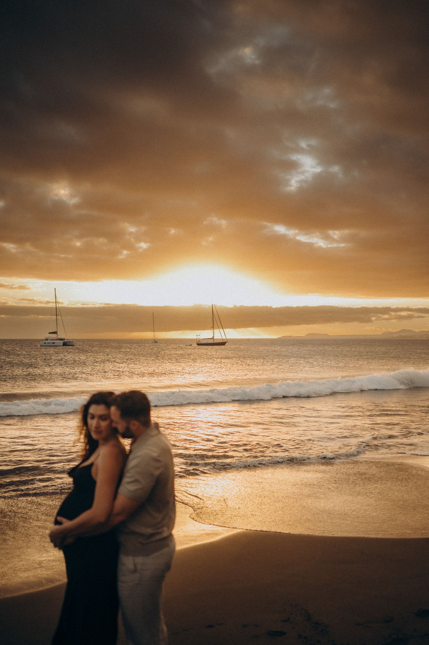 Couple embracing on a sandy beach at golden sunset with sailboats on the ocean horizon.