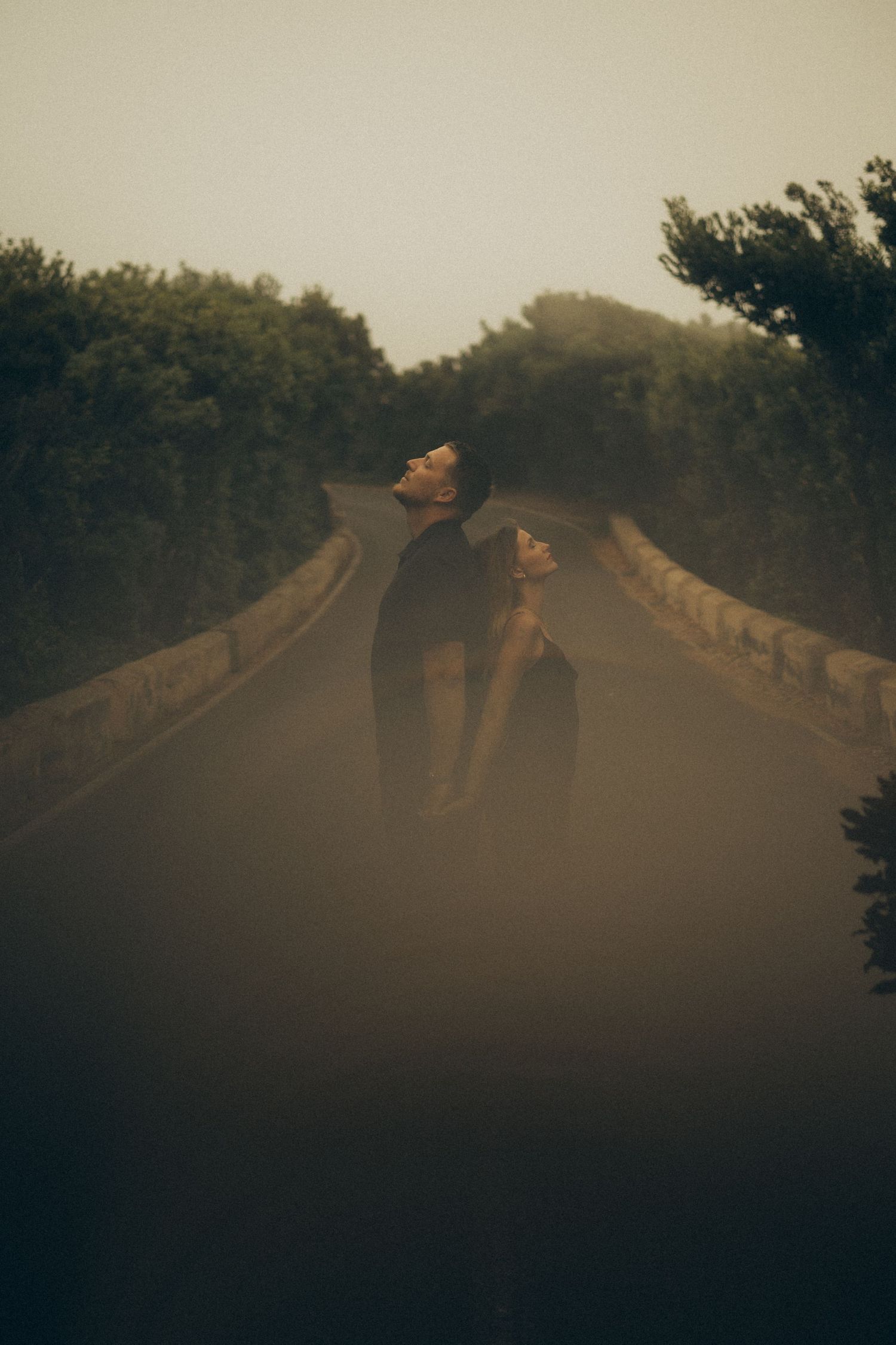 Two people stand embracing on a misty road surrounded by trees in a moody, foggy atmosphere.