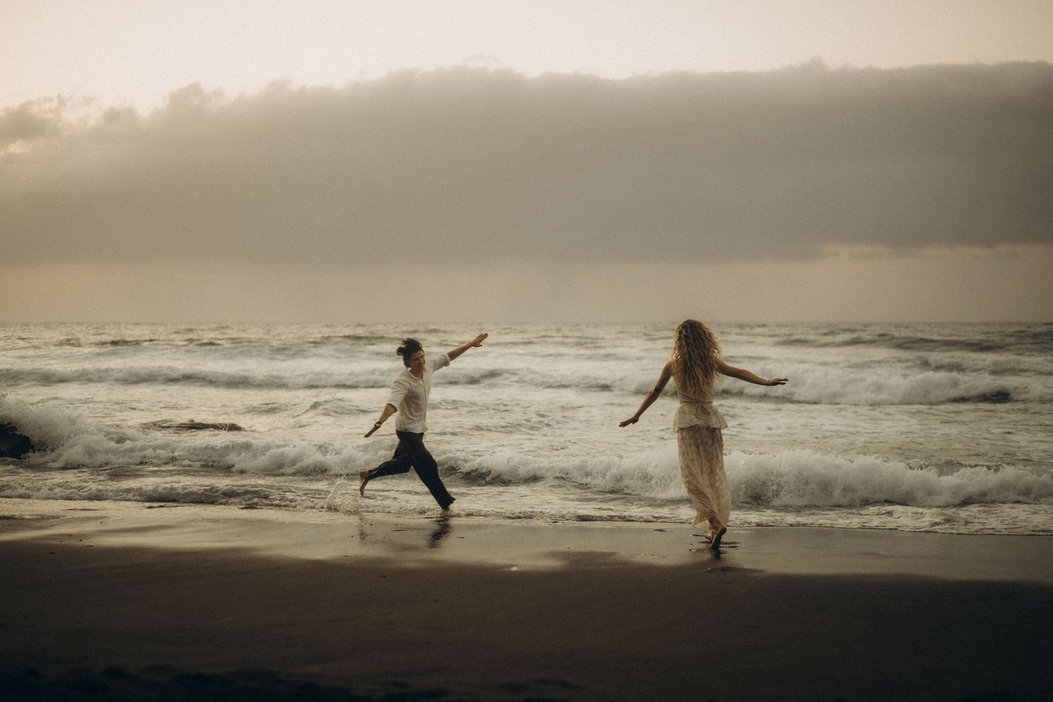 Couple running playfully along a misty beach at sunset with ocean waves crashing behind them.