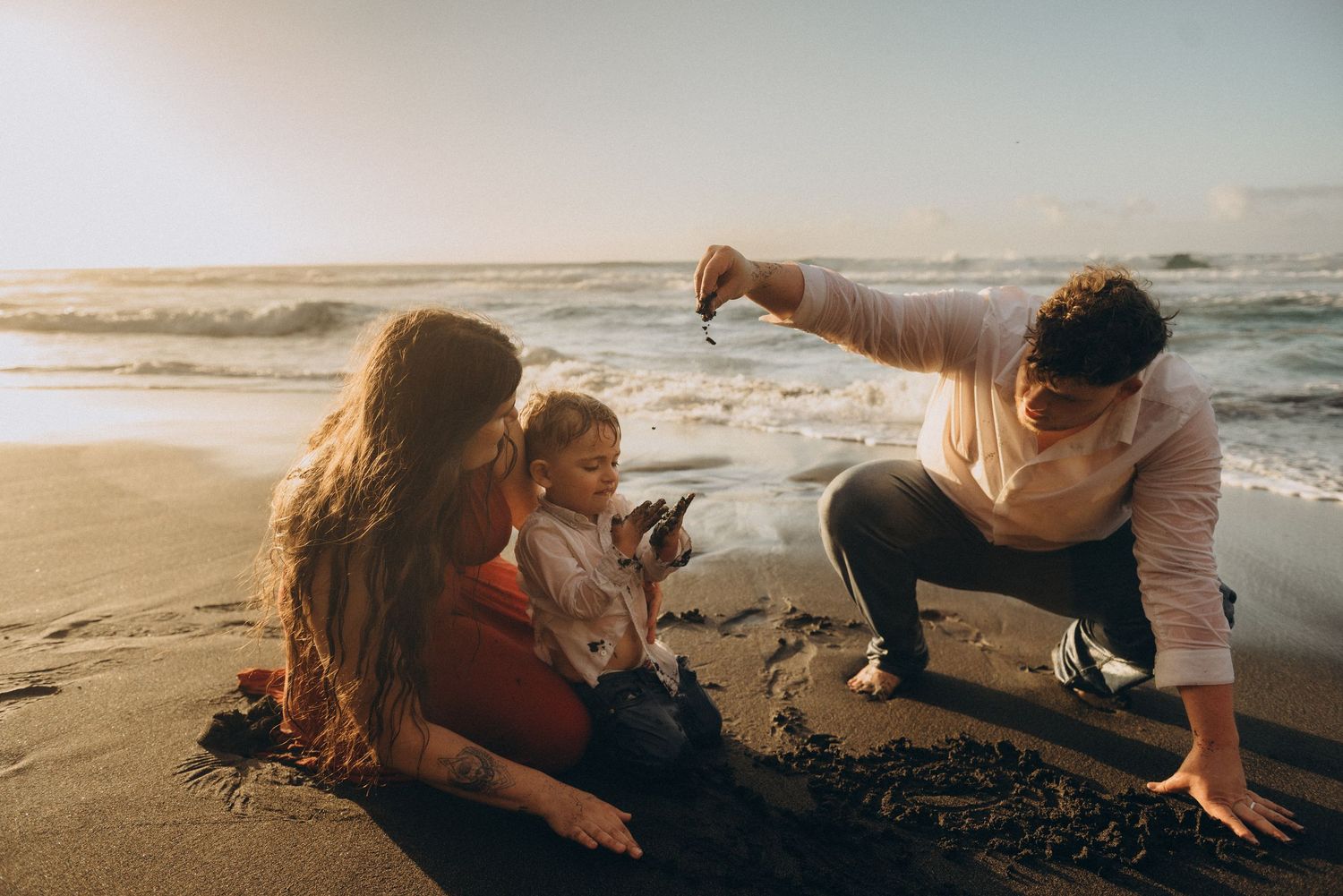 Family enjoying a beach sunset, with a child sitting on wet sand as a parent holds up a small object.