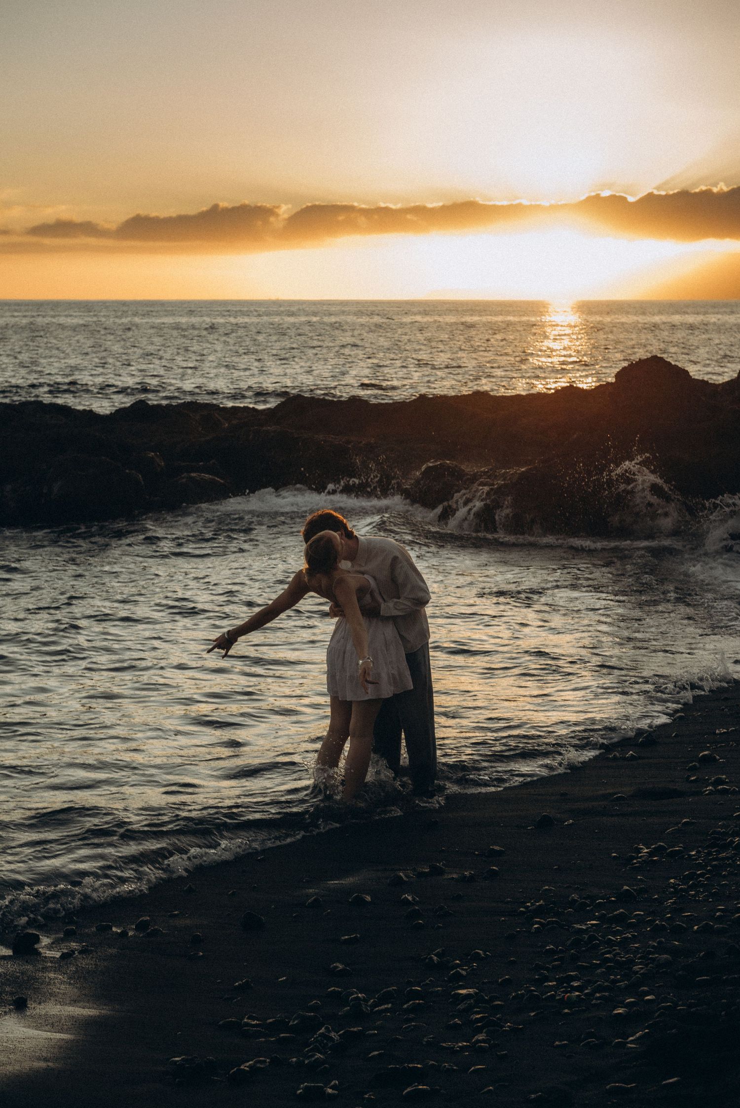Couple embracing on a black sand beach at golden sunset, waves gently washing over their feet near rocky shore.