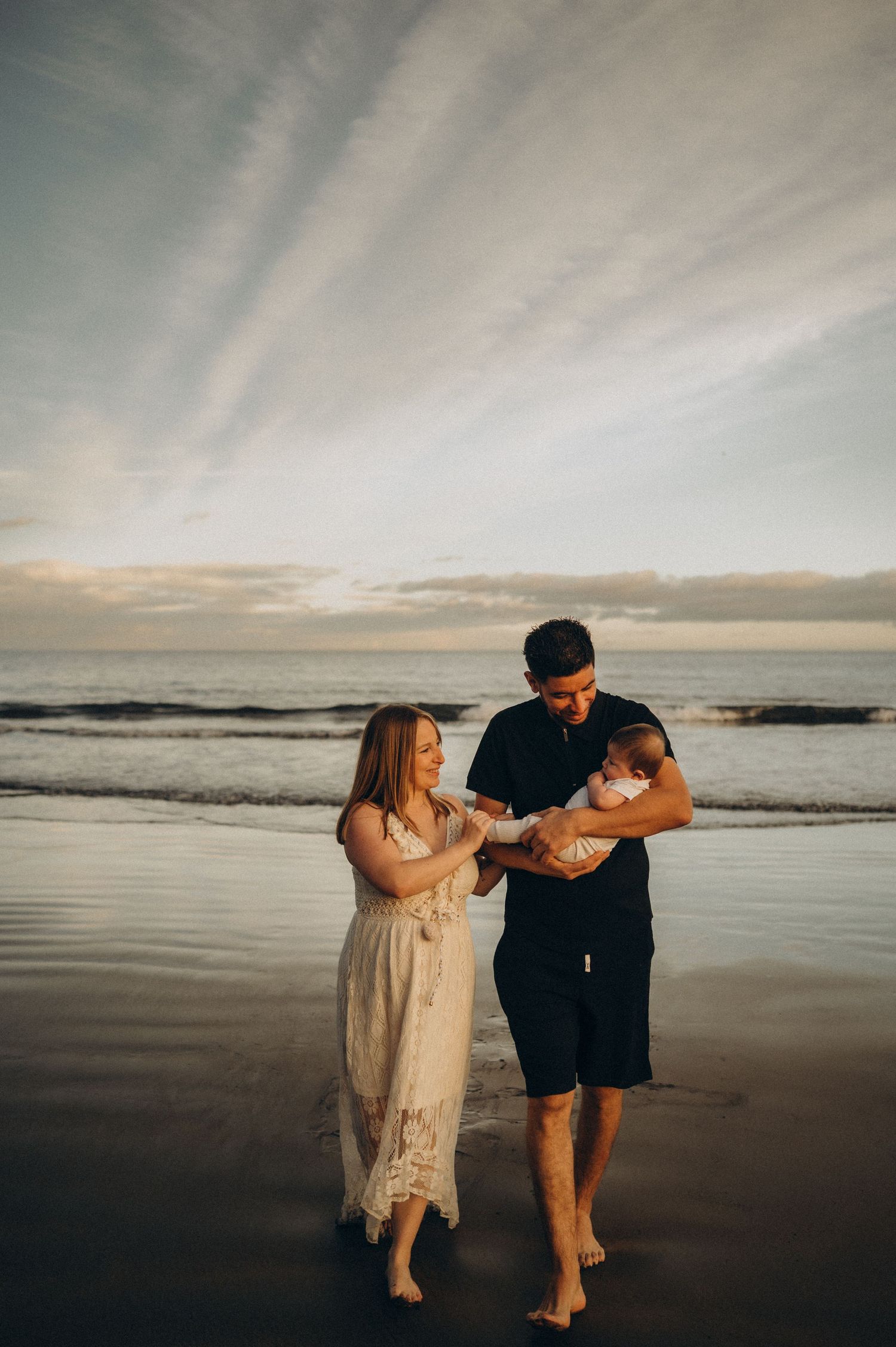 Family holding baby dancing together on moody Tenerife beach at sunset with romantic silhouettes and ocean waves during photoshoot.