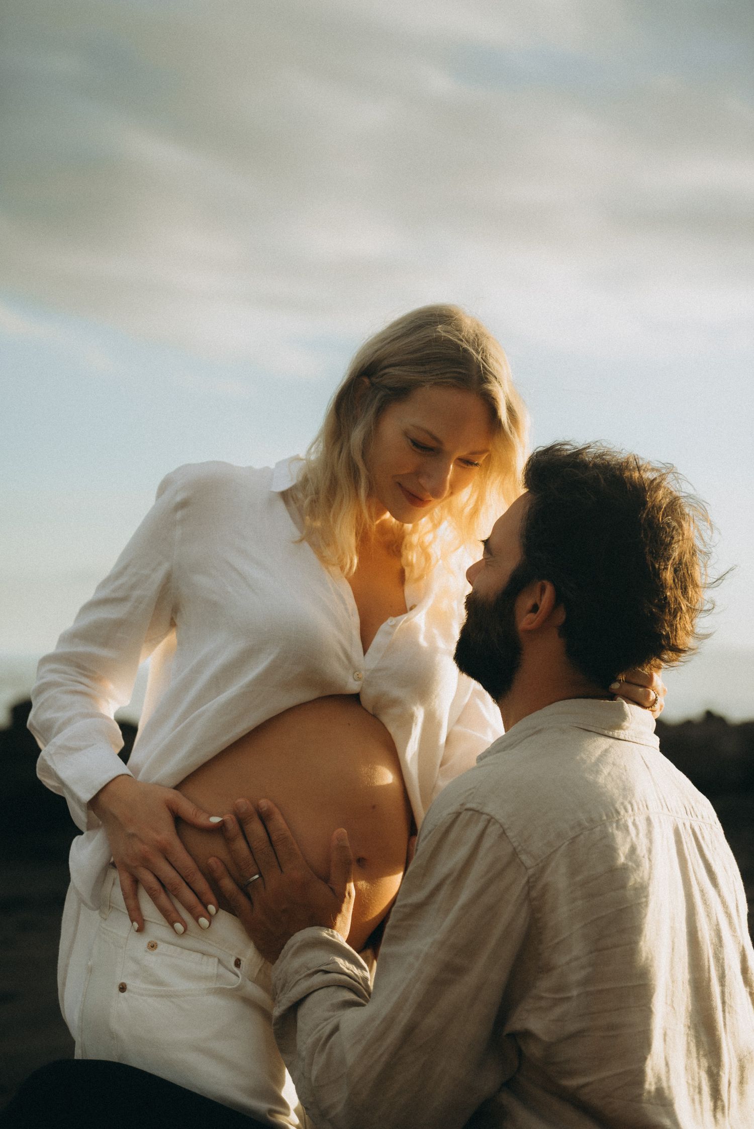Pregnant woman in white shirt with partner touching her bare belly during golden hour outdoor maternity photoshoot.