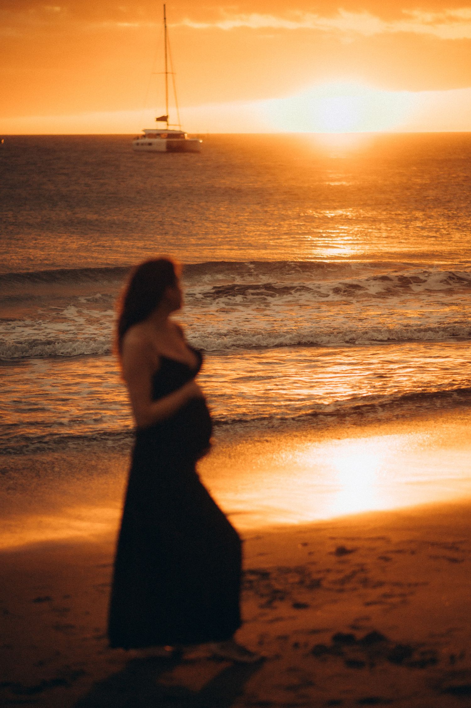 Pregnant woman in black dress standing on beach at golden sunset with sailboat on horizon.