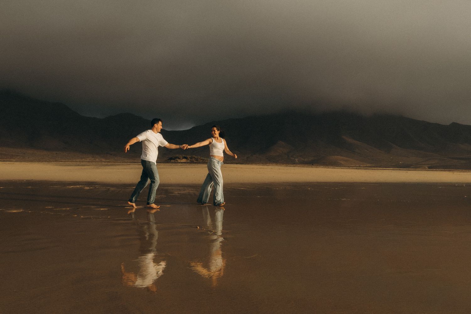 Couple dancing on a reflective beach with dramatic storm clouds and mountains in the background at golden hour.