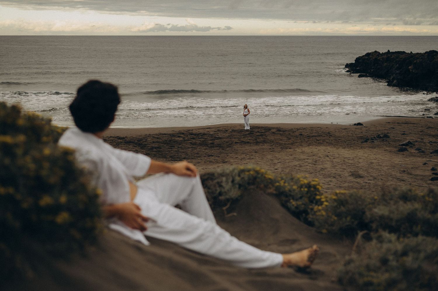 A person in white sits on a sandy cliff overlooking a beach where another figure stands by the ocean.