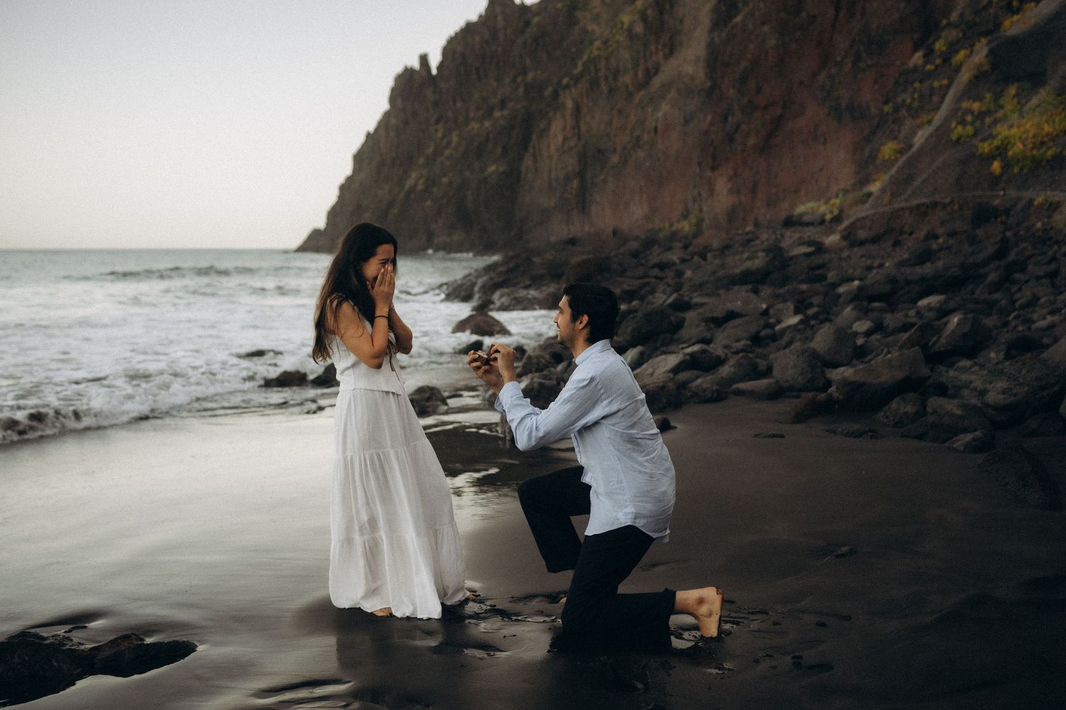Surprise proposal in Tenerife at Playa de las Gaviotas on a rocky black sand beach at sunset with a joyful reaction of surprise.