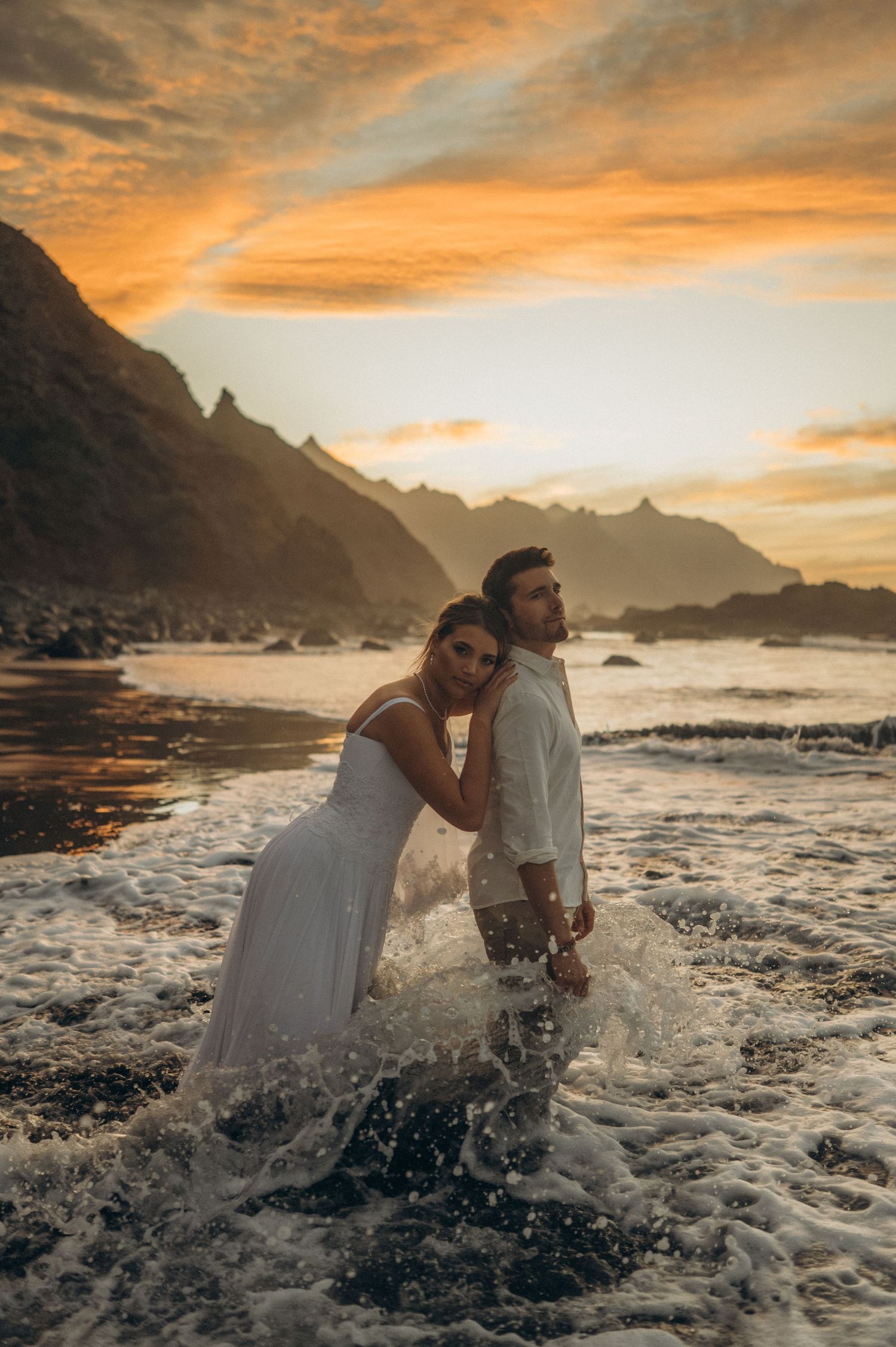 Bride hugging groom in the water with majestic cliffs and orange-pink sunset at Playa Benijo, Tenerife