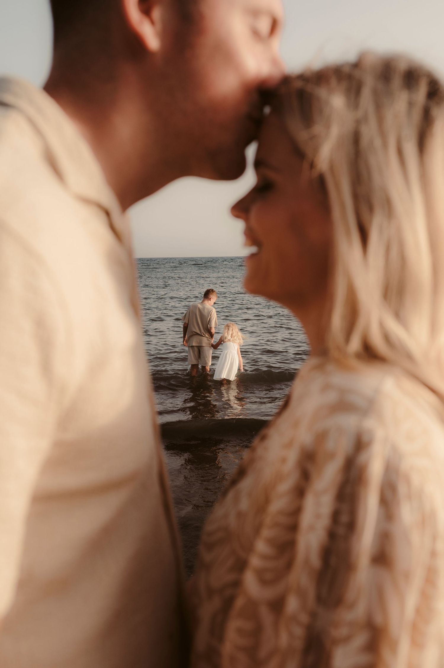 Close-up of man kissing partner’s forehead with children playing in ocean behind