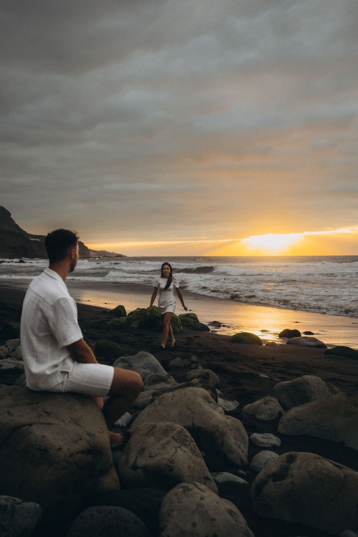 Couple on rocky beach at sunset, man sitting on rocks watching woman in white dress near ocean waves under dramatic cloudy sky.