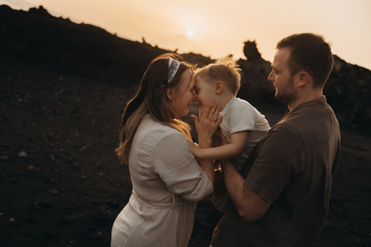 A family shares a tender moment at sunset, with parents kissing their toddler against a dramatic rocky landscape.