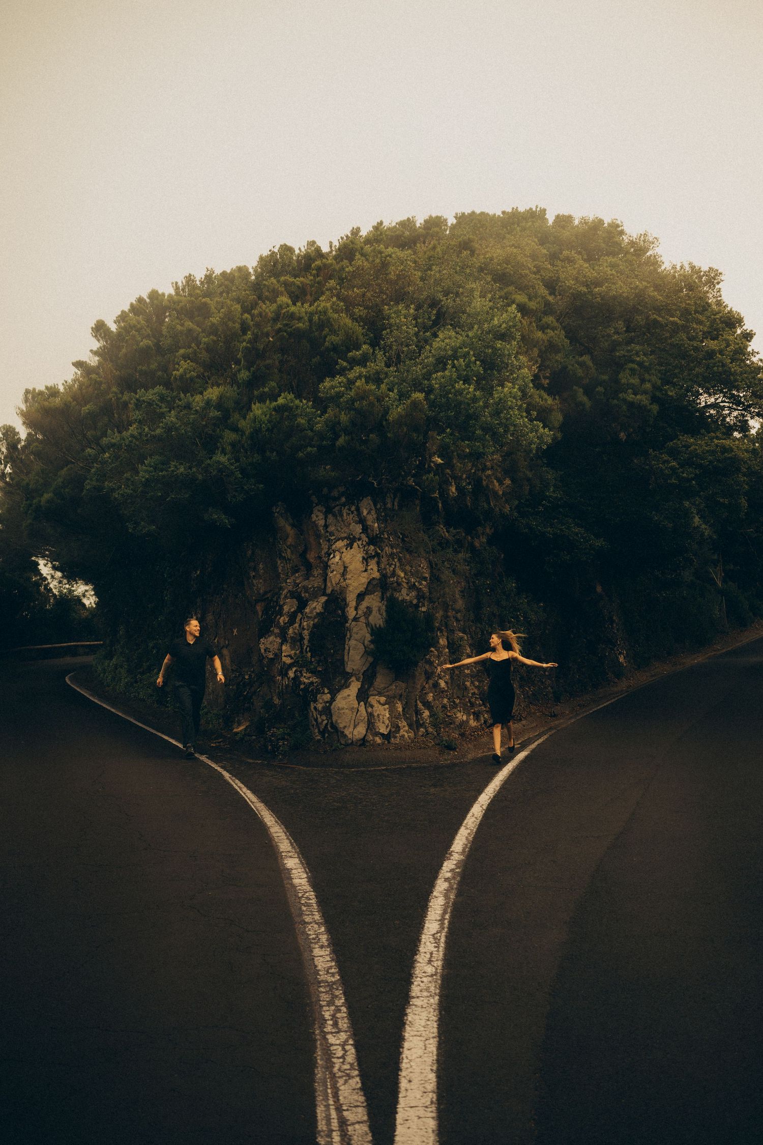 Two people stand at a forked road diverging around a rocky tree-covered hill at dusk.