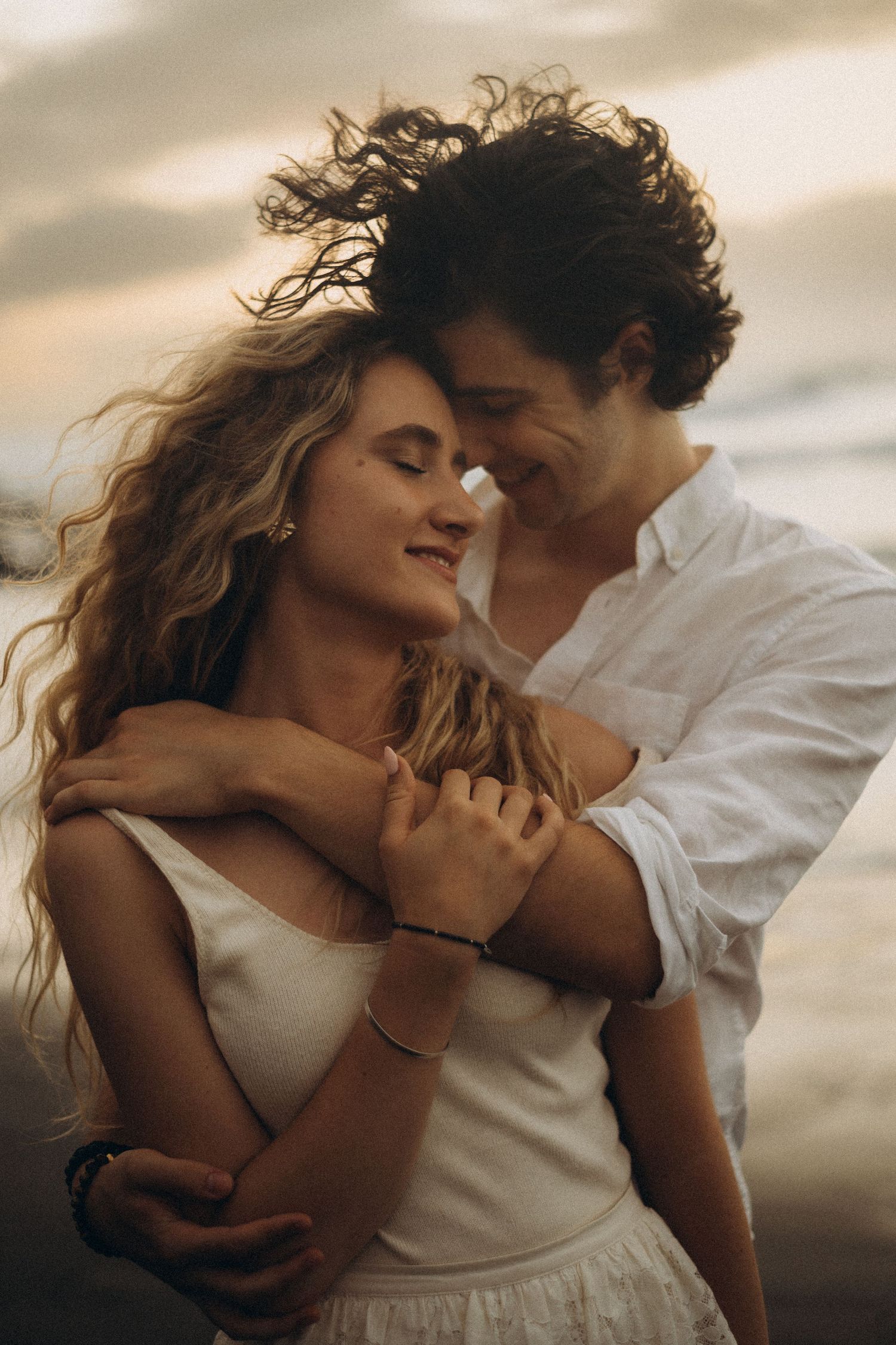 Romantic couple embracing on a windy beach at sunset, woman in white dress with flowing hair, man in white shirt.