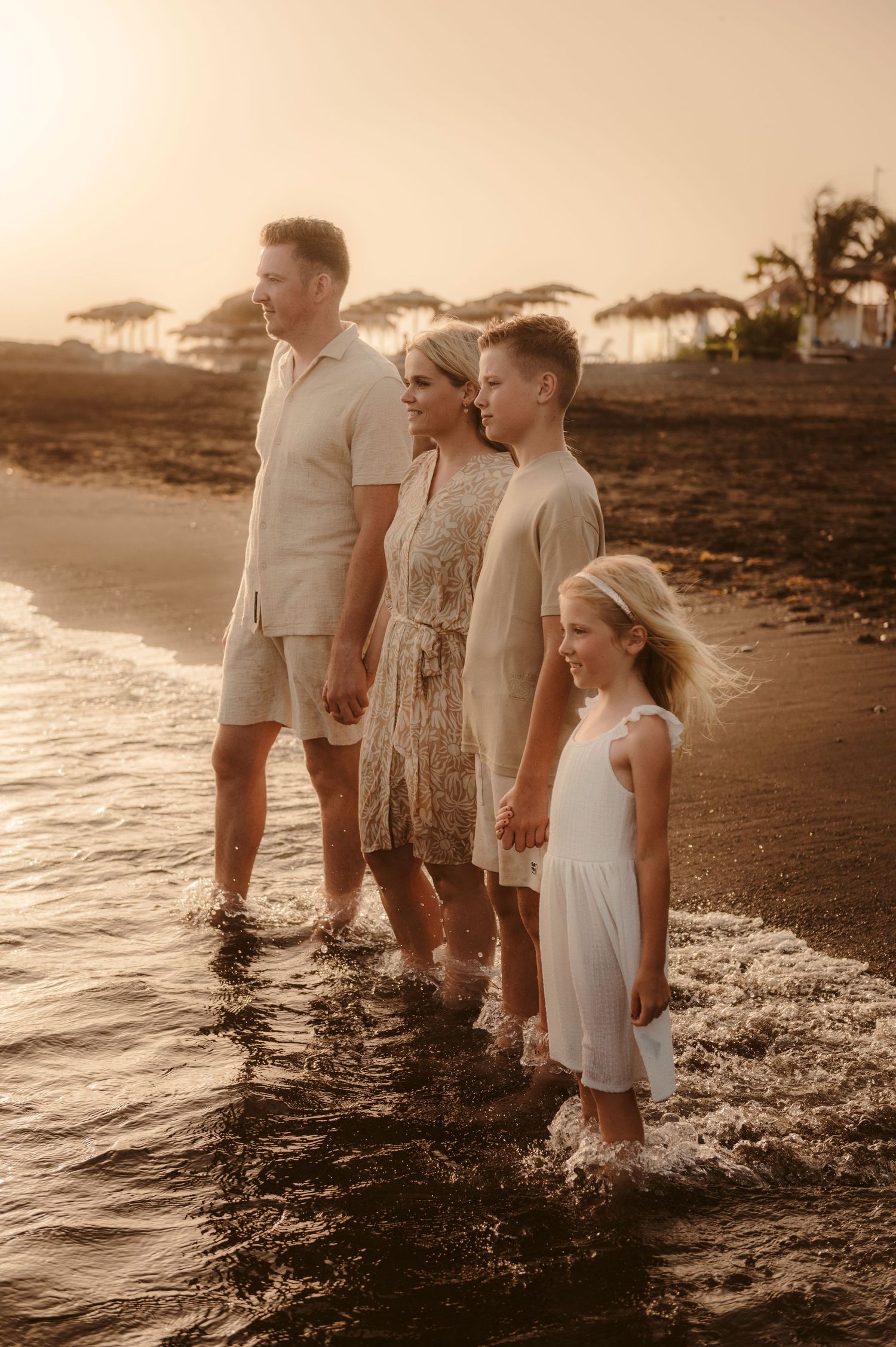 Family holding hands in a line standing on beach with feet in ocean at golden hour, Playa del Duque.