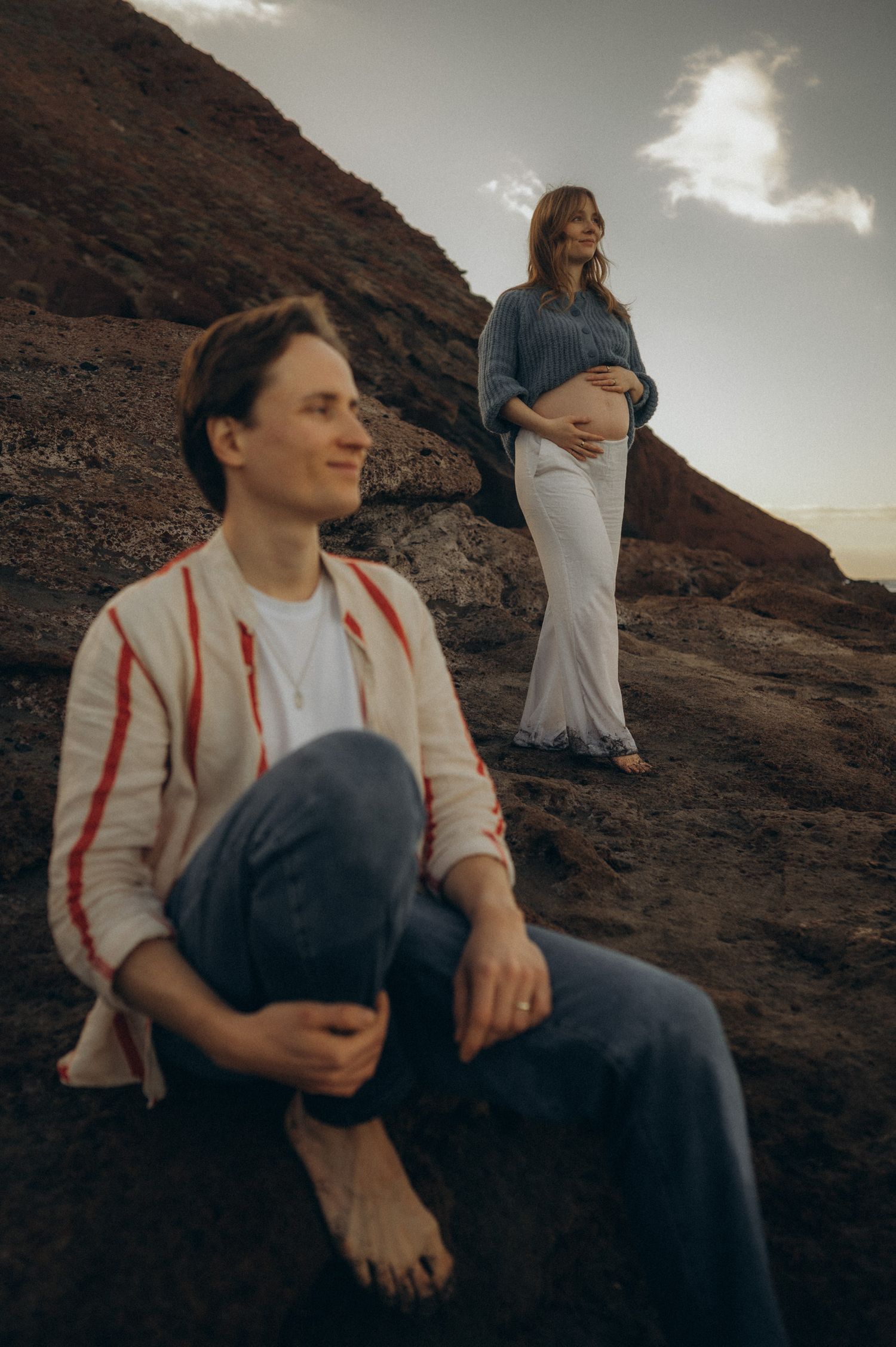 A barefoot man sits on rocky terrain while a pregnant woman stands behind him holding her belly outdoors.