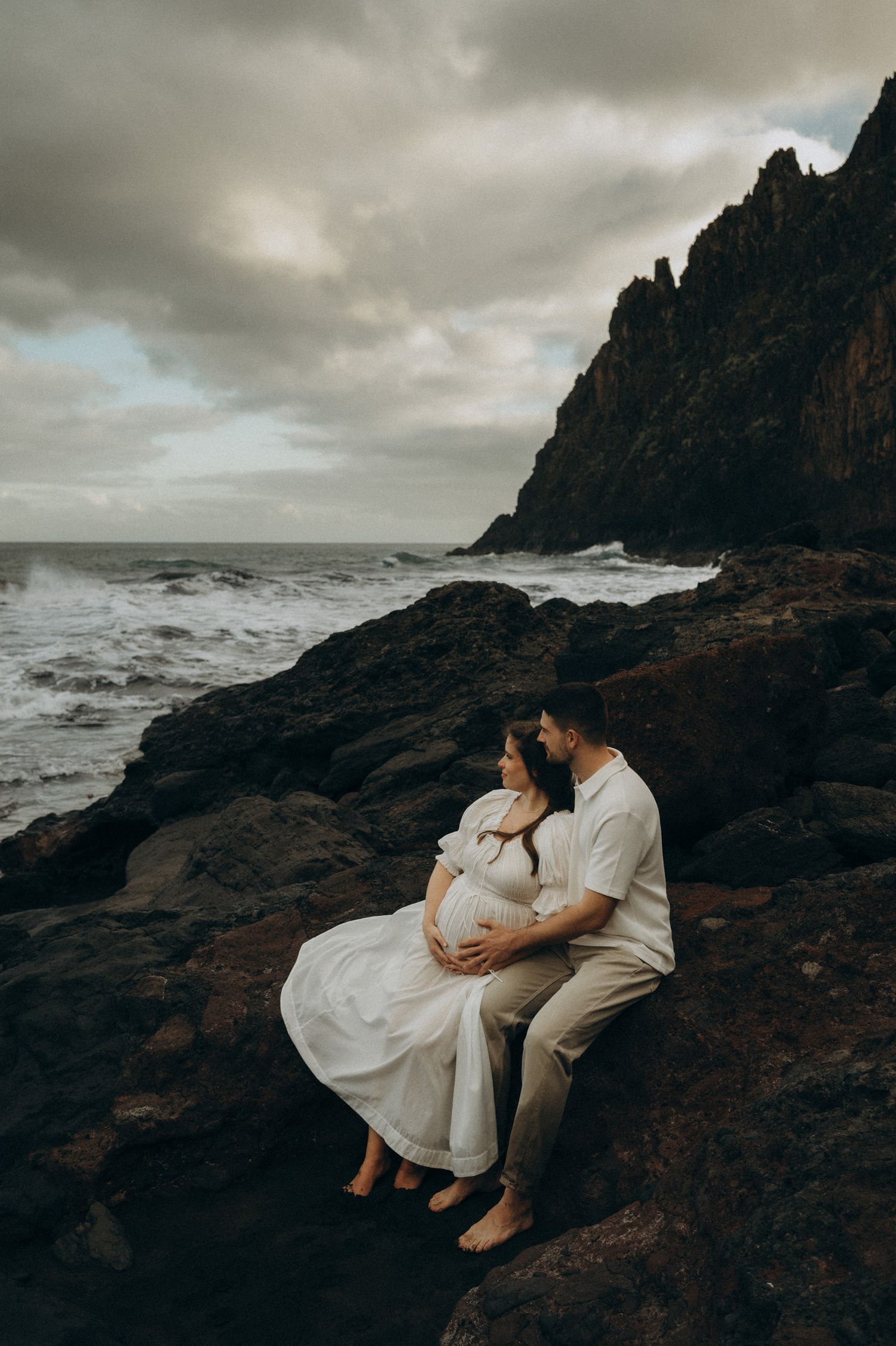 Couple sharing a tender maternity moment on rocky coastal cliffs with dramatic waves and stormy skies.