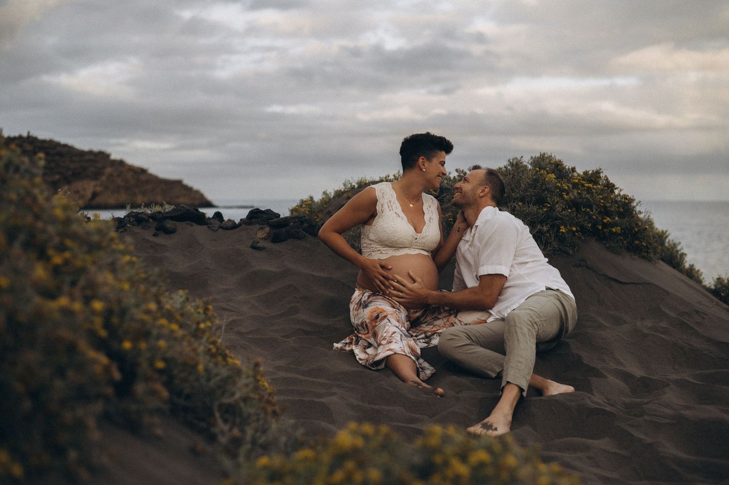 Couple sitting on dark sand dunes by the ocean, sharing a tender moment during a maternity photoshoot.