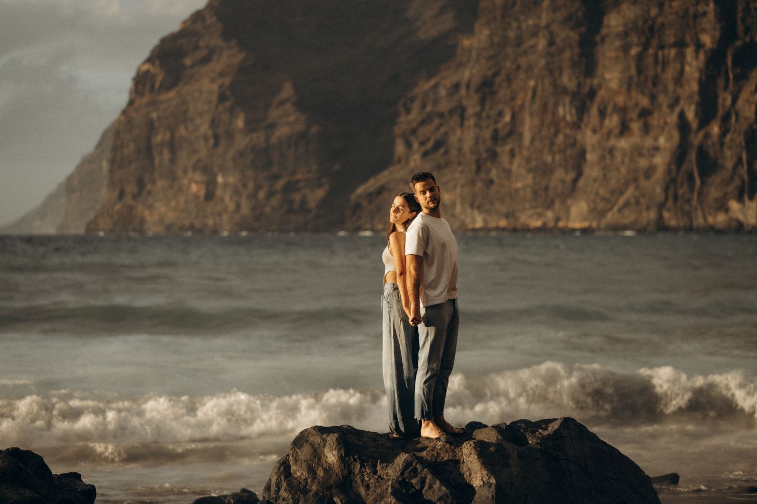 Couple standing back-to-back in embrace on Playa de Los Guíos with Los Gigantes cliffs, Tenerife