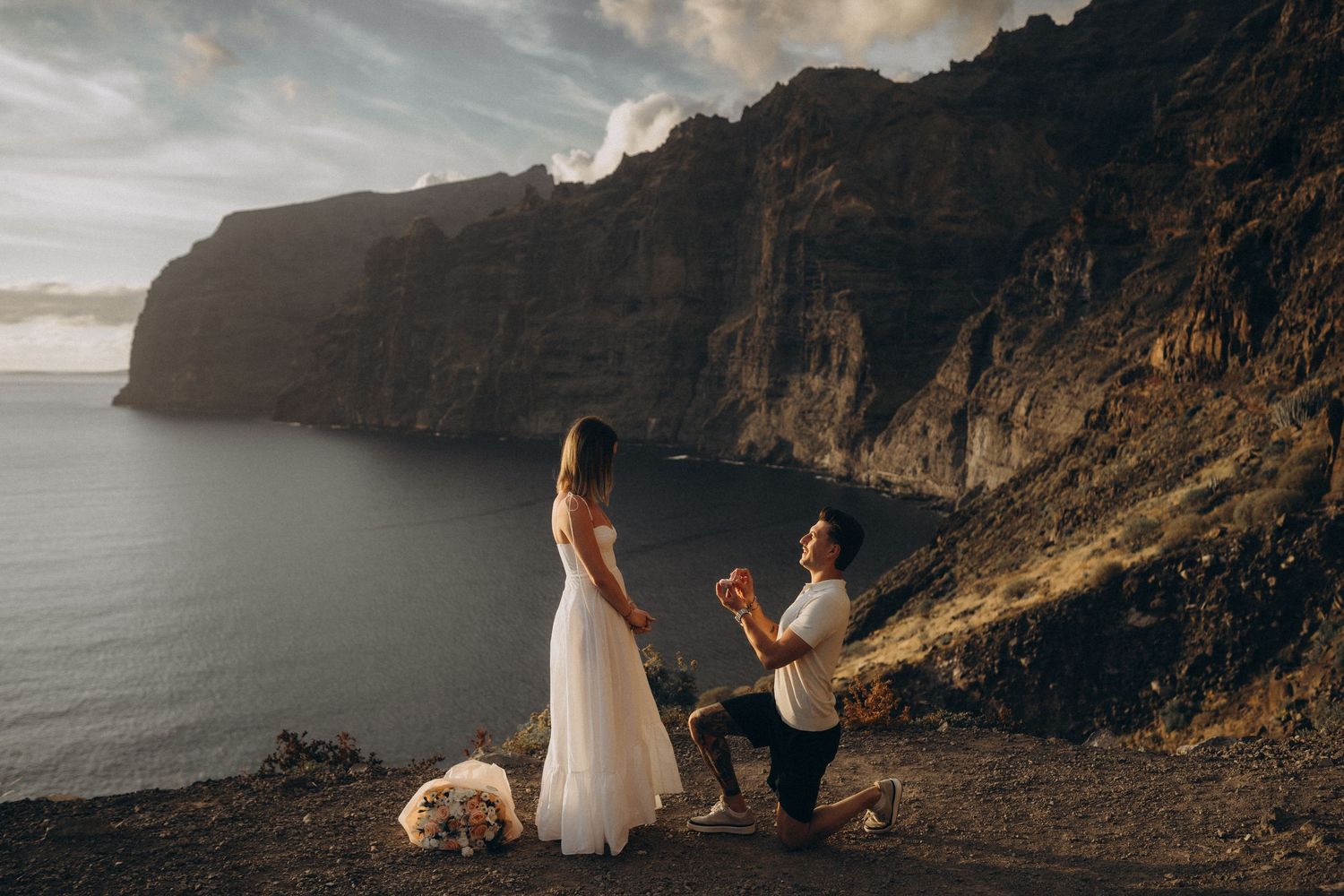 Man proposes to woman in white dress on cliffside overlooking ocean with dramatic mountain backdrop at sunset.