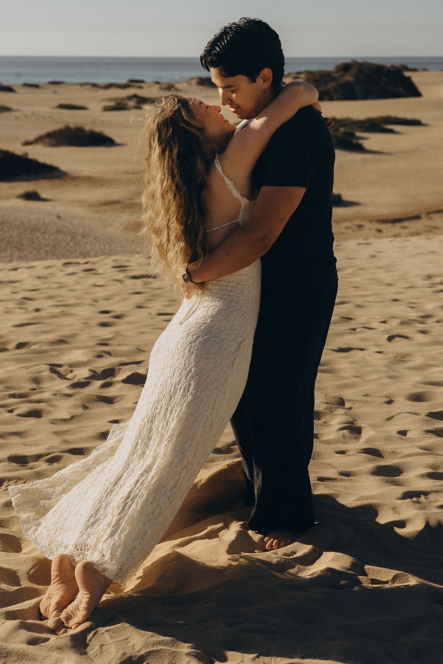 Couple embracing romantically on sandy beach dunes, woman in white lace dress, man in black, ocean in background.