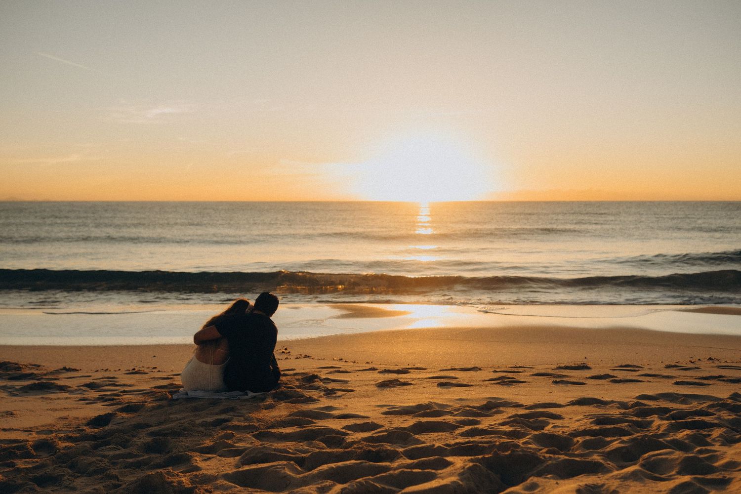 Couple sitting together on a sandy beach watching a golden sunset over the ocean waves.