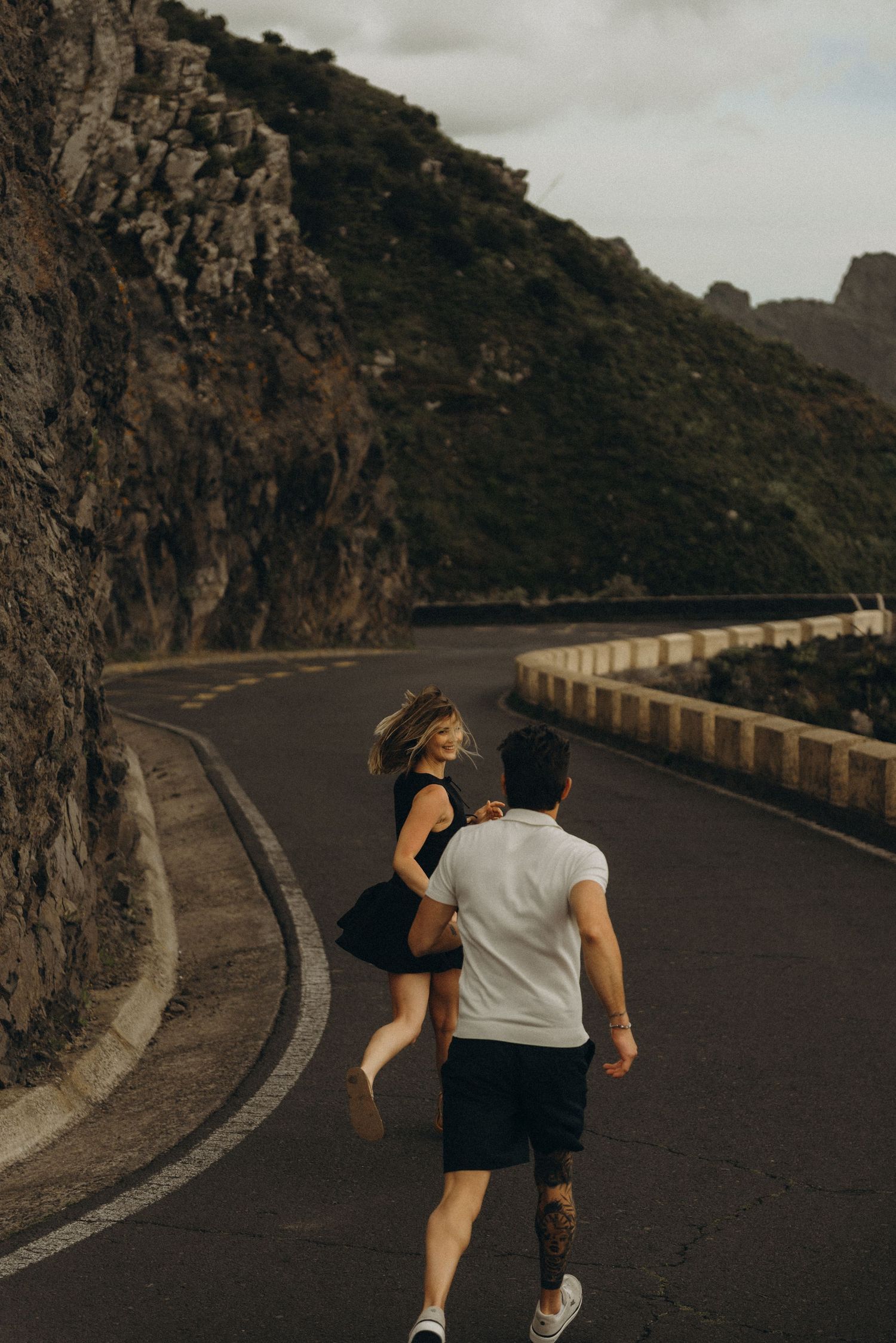 Couple running together on a winding mountain road, woman in black dress and man in white shirt with tattoos.