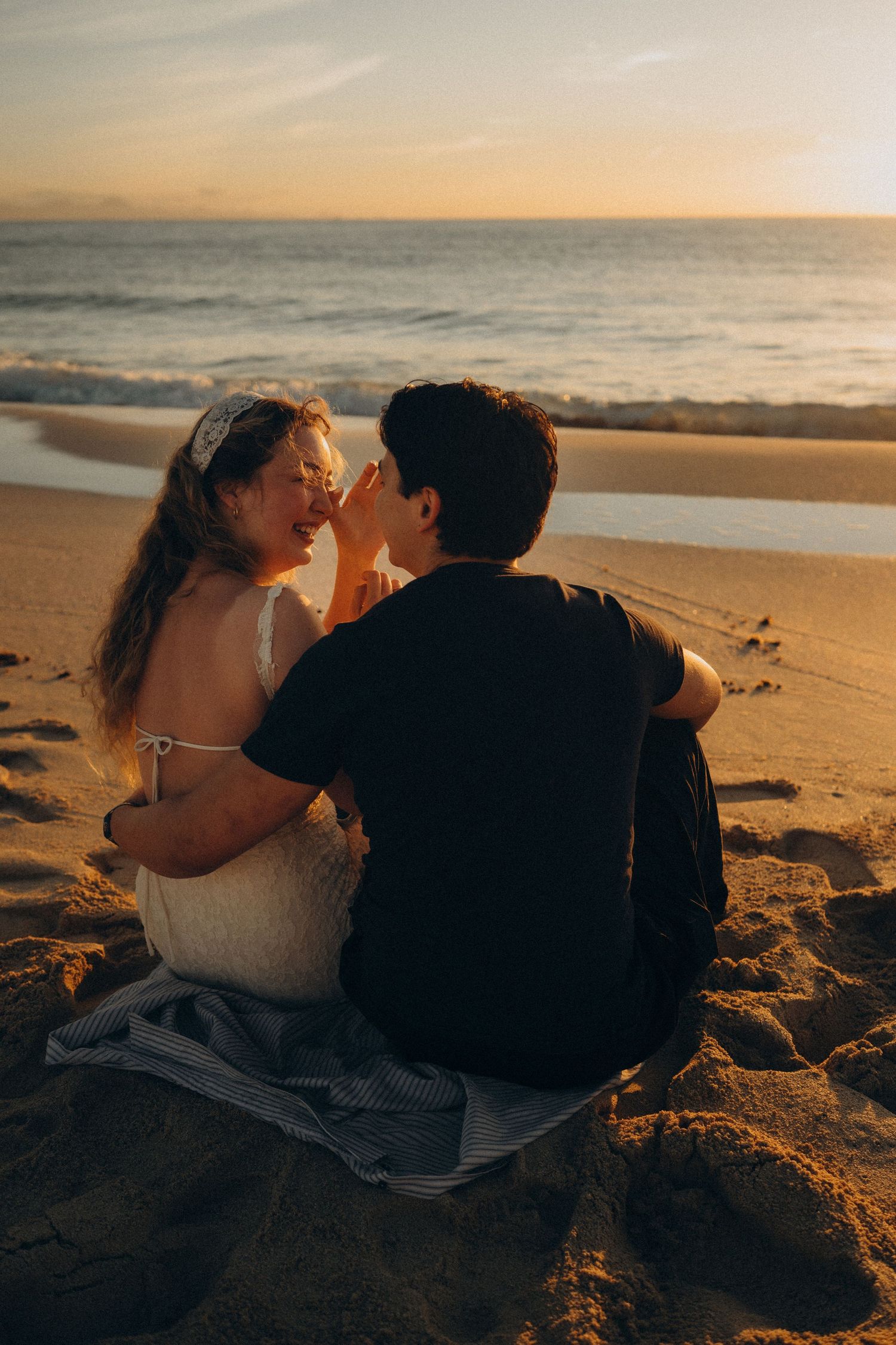 Couple sitting on beach blanket at sunset, sharing a romantic moment by the ocean waves.