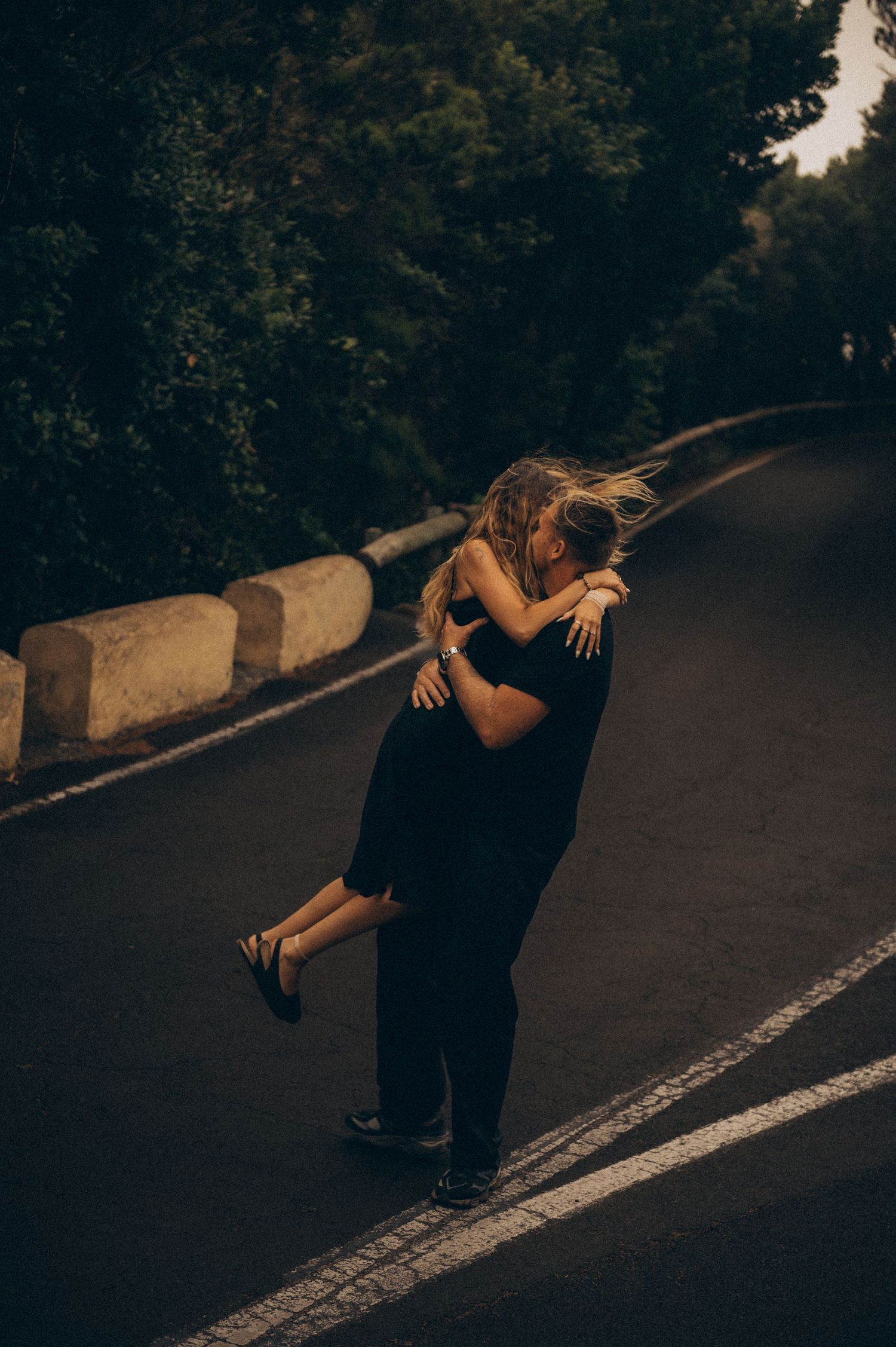 Couple embracing and kissing on a dark road surrounded by lush green trees, wearing elegant black outfits.