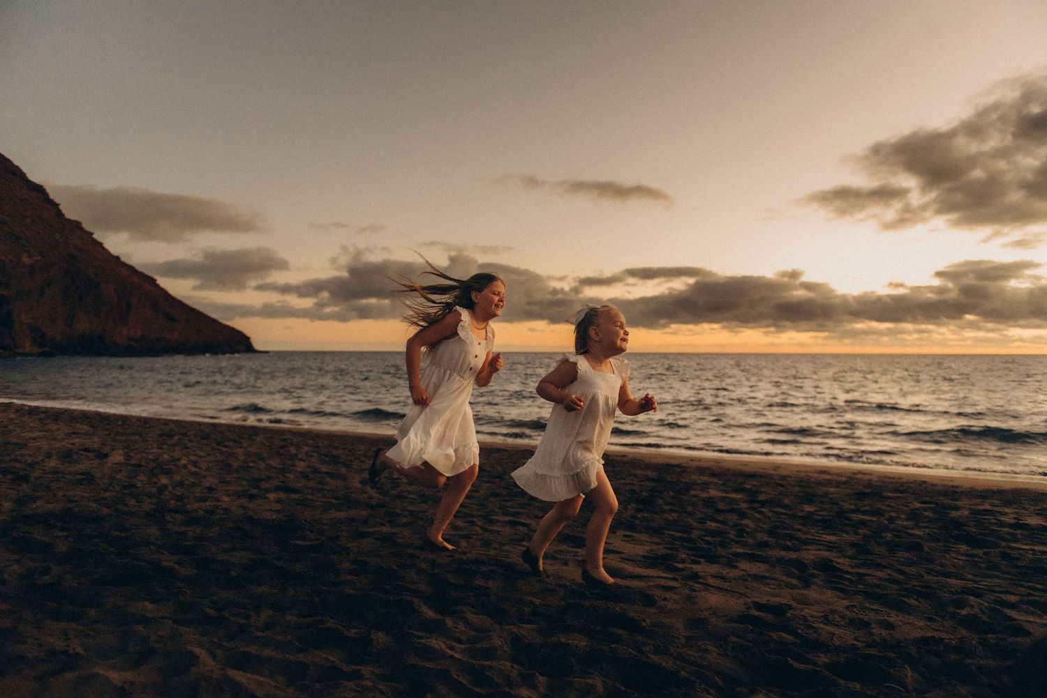 Family photoshoot on black sand beach in Tenerife with two children running, two people in white outfits walking, and dramatic sunset clouds