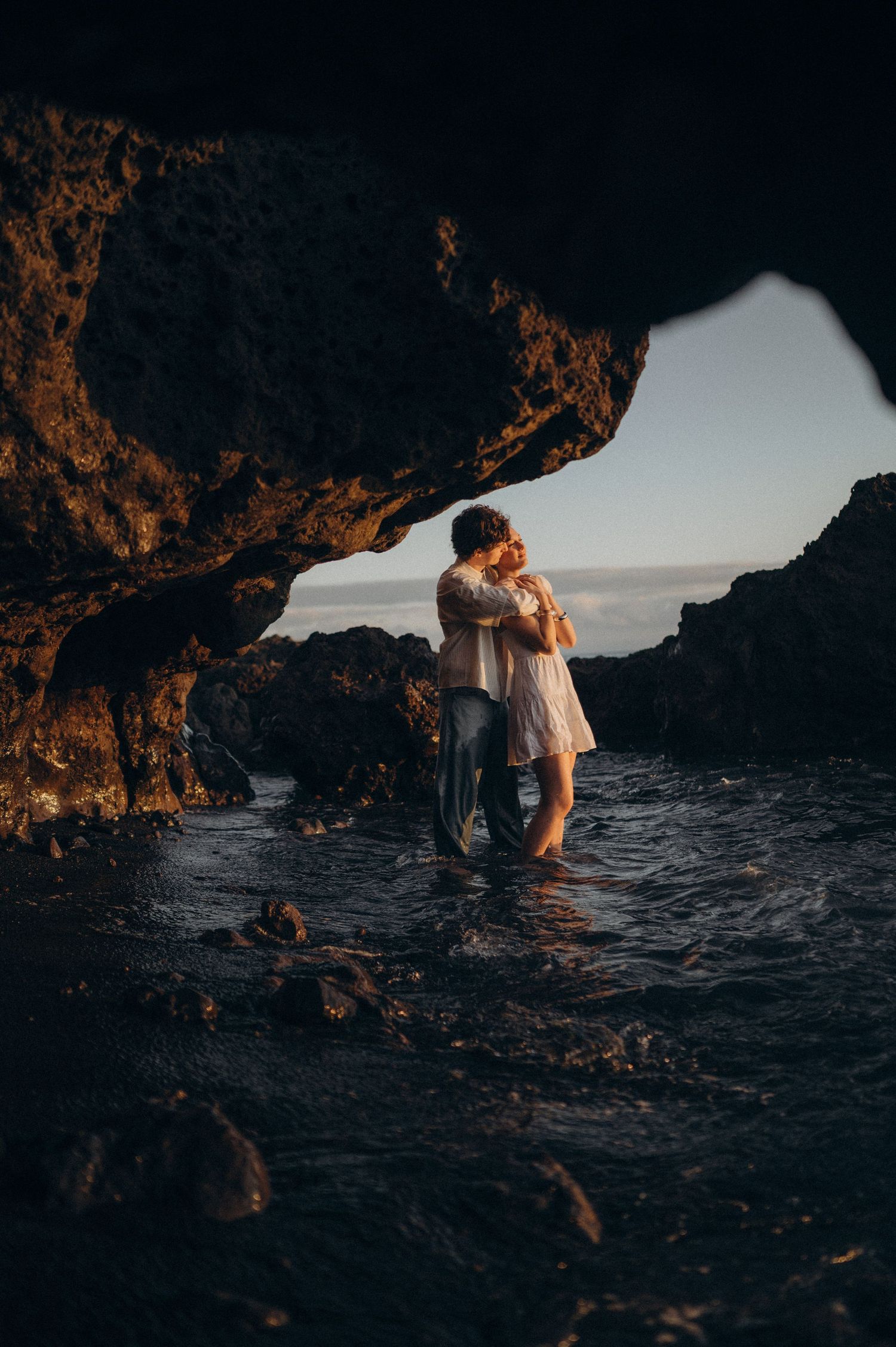 Couple embracing romantically in a sea cave at golden hour, standing in shallow water with dramatic rock formations.