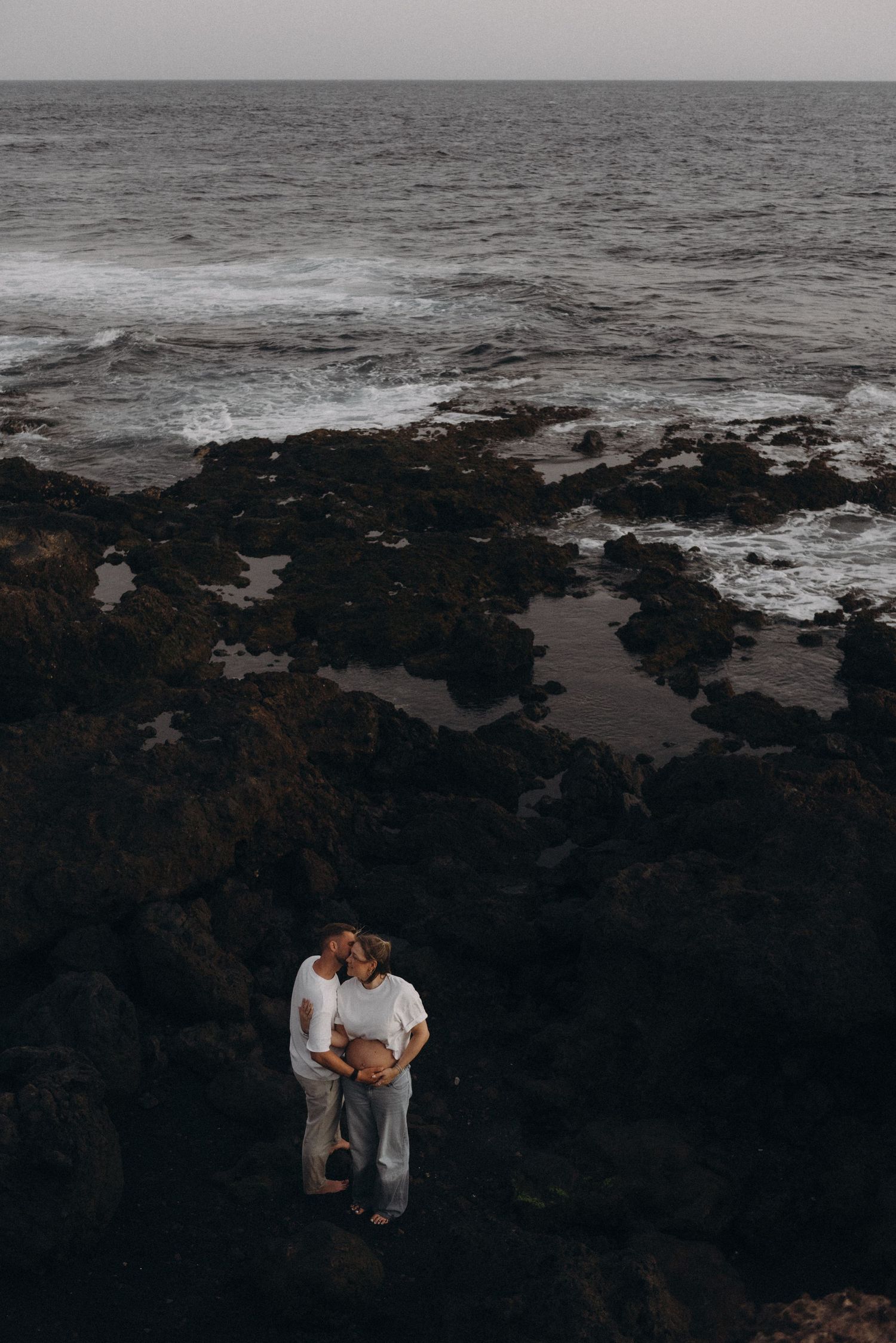 Couple embracing on dark volcanic rocks by the ocean shore, sharing a tender moment with waves in the background.