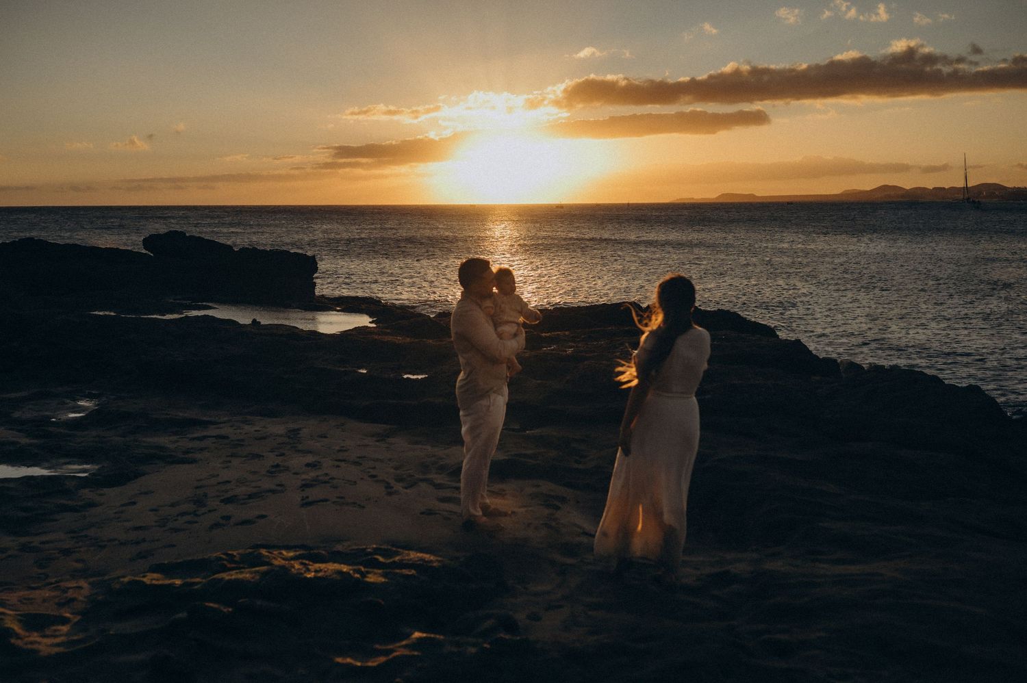 A series of silhouetted figures stand on a rocky coastline during a dramatic golden sunset over the ocean.