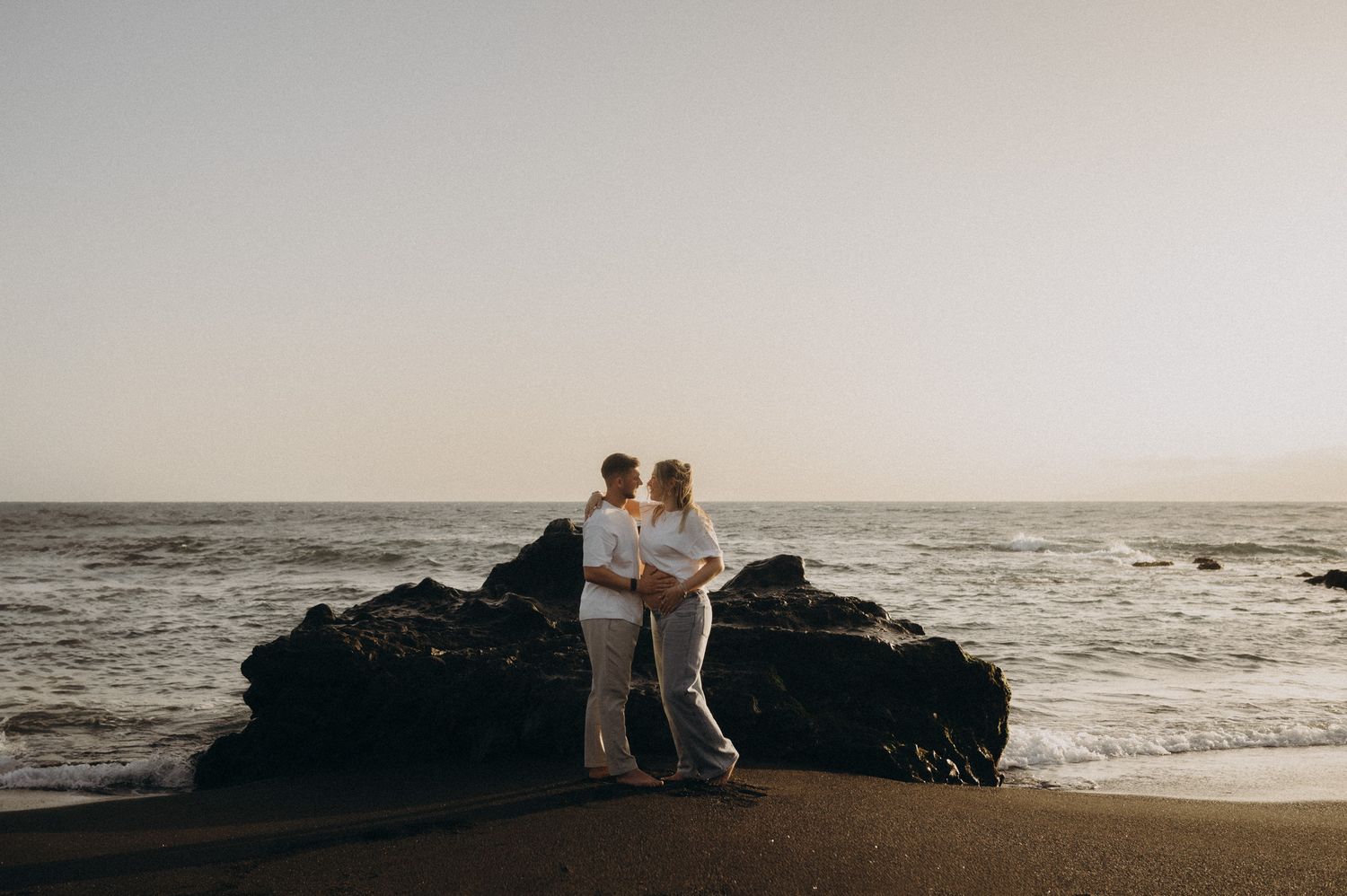 Couple embracing on a rocky beach at sunset with ocean waves and golden sky in the background.