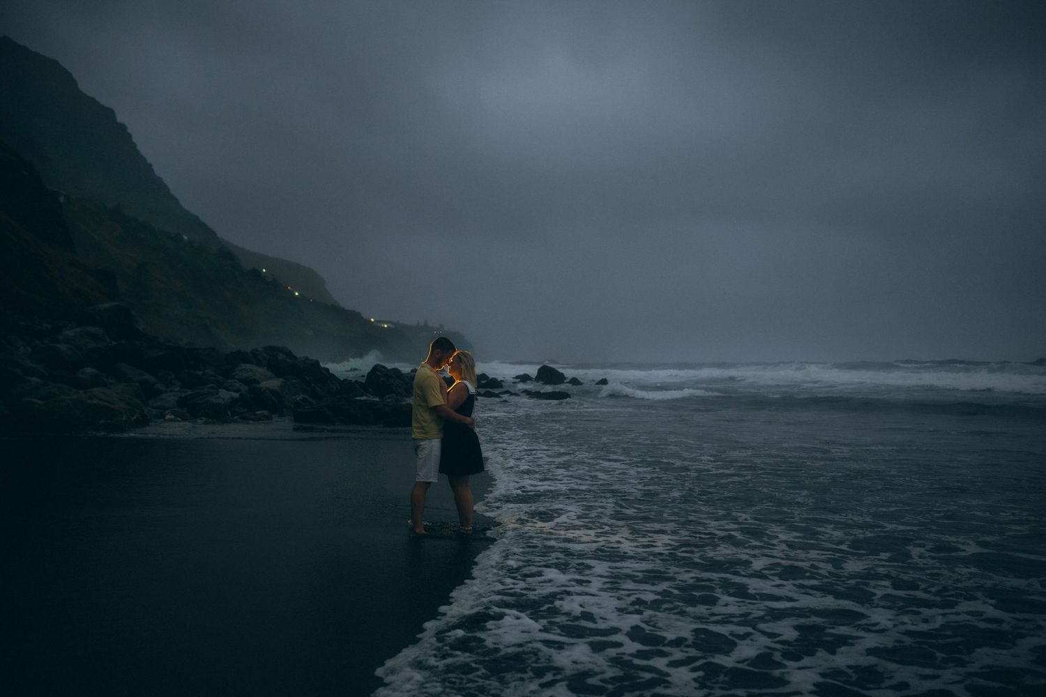 Couple embracing on a dark moody beach at dusk with dramatic cloudy skies and rocky cliffs in background.
