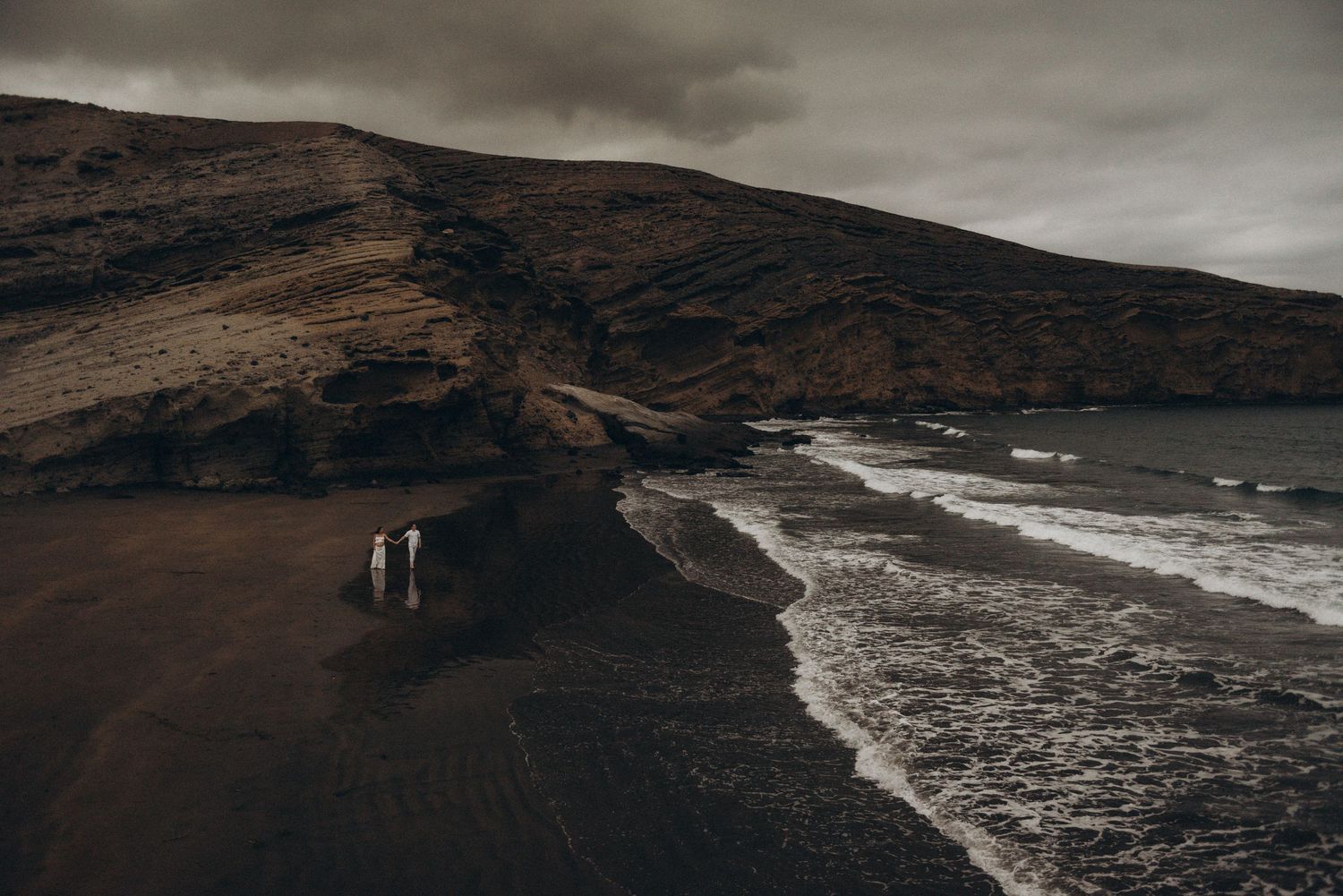 Couple standing on a dramatic dark volcanic black sand beach with moody stormy skies and rugged cliffs.
