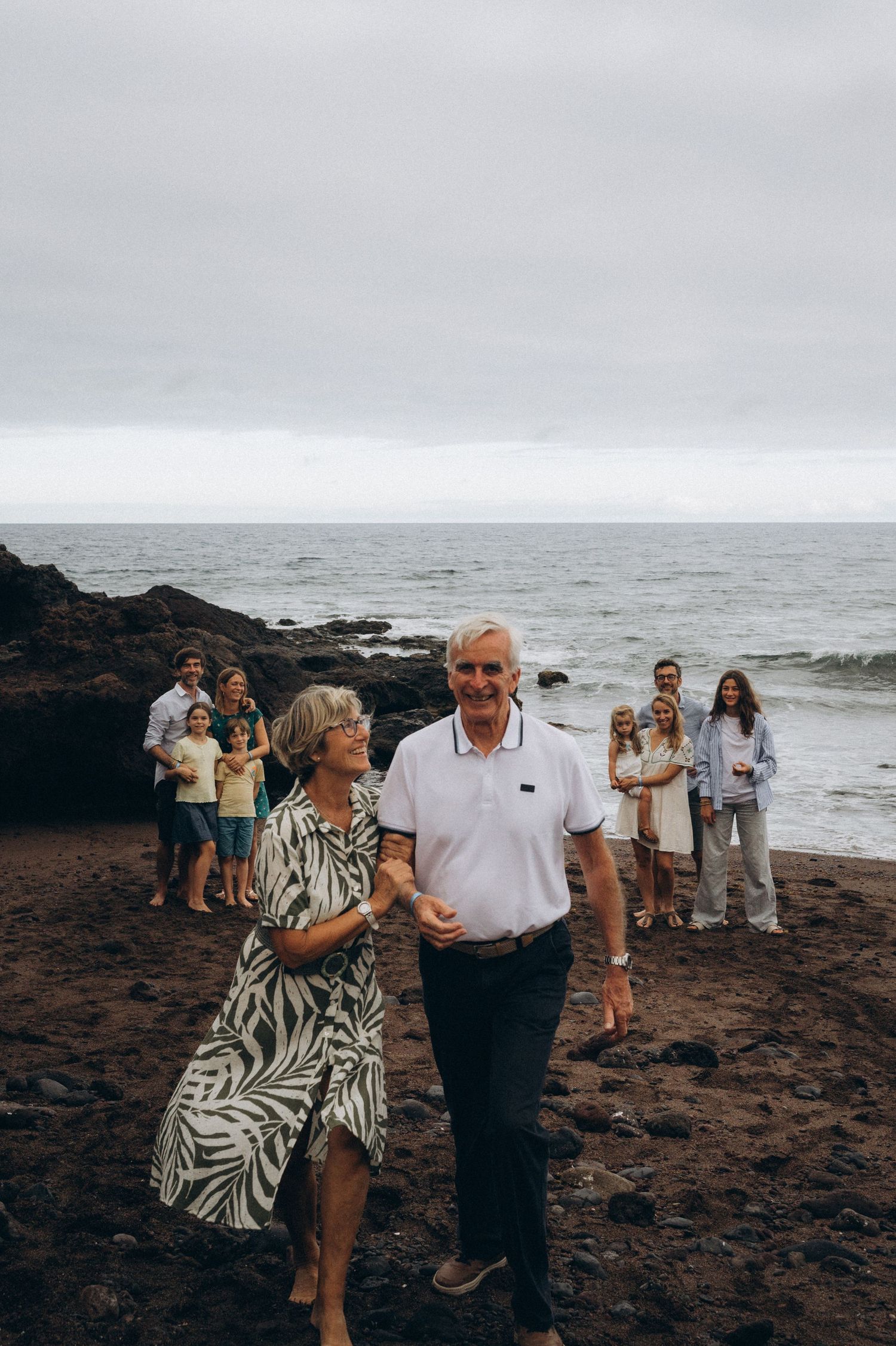 Two fathers standing with their families on black sand as grandparents walk toward camera at Los Refugios